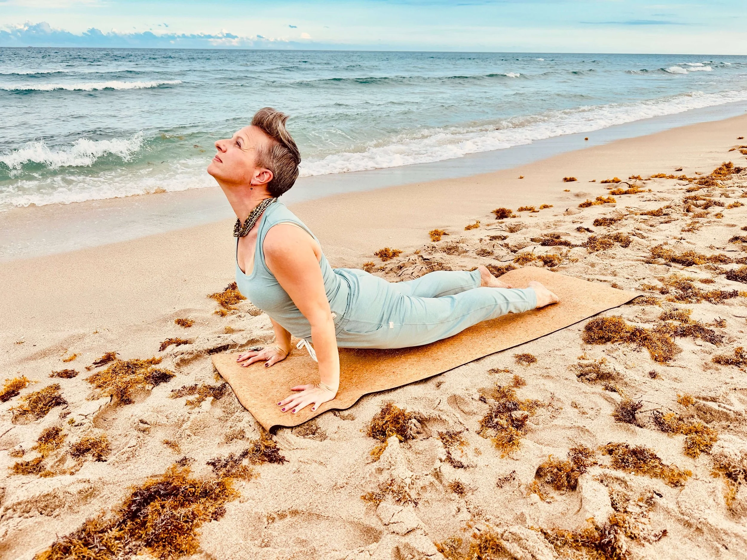 Angela Towne practicing yoga on the beach, in a cobra pose with elbows bent, looking upwards, with ocean waves and cloudy sky in the background.