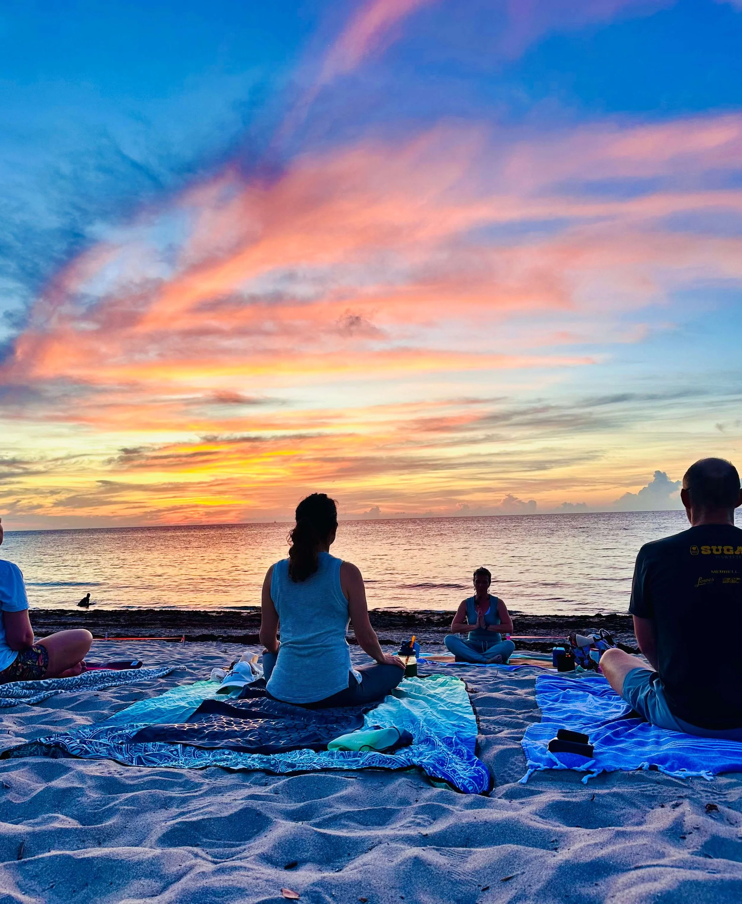 Group of people practicing yoga on a beach during sunset with colorful sky and calm ocean.