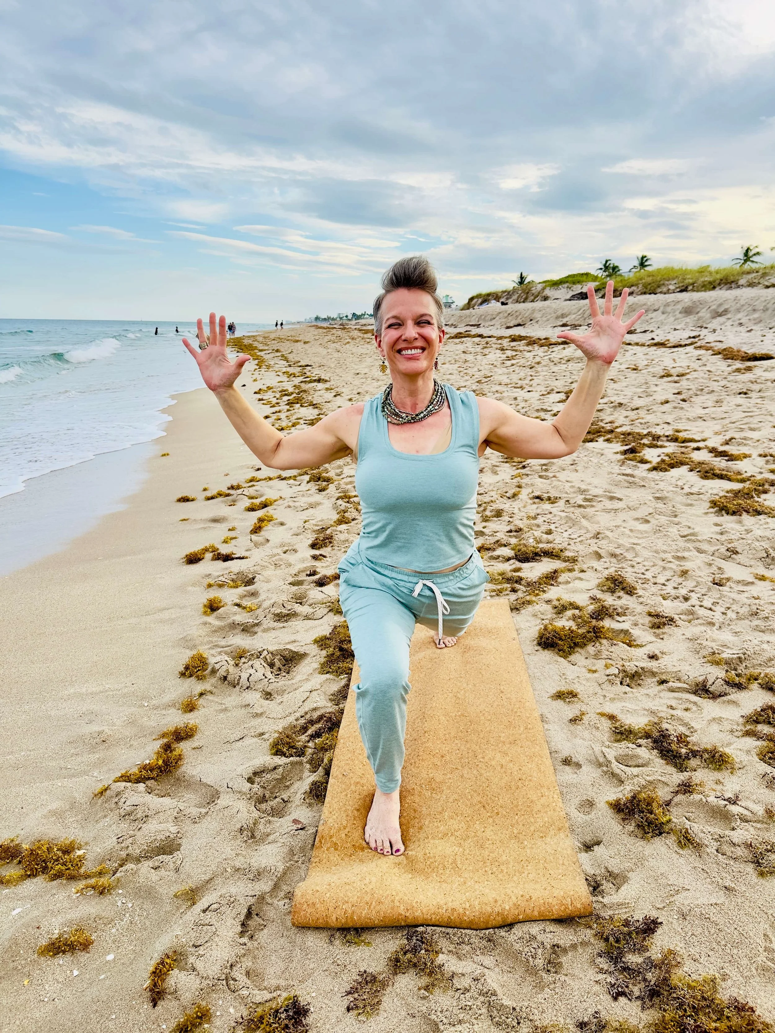 A woman practicing yoga on a sandy beach, smiling and raising her arms with palms open.