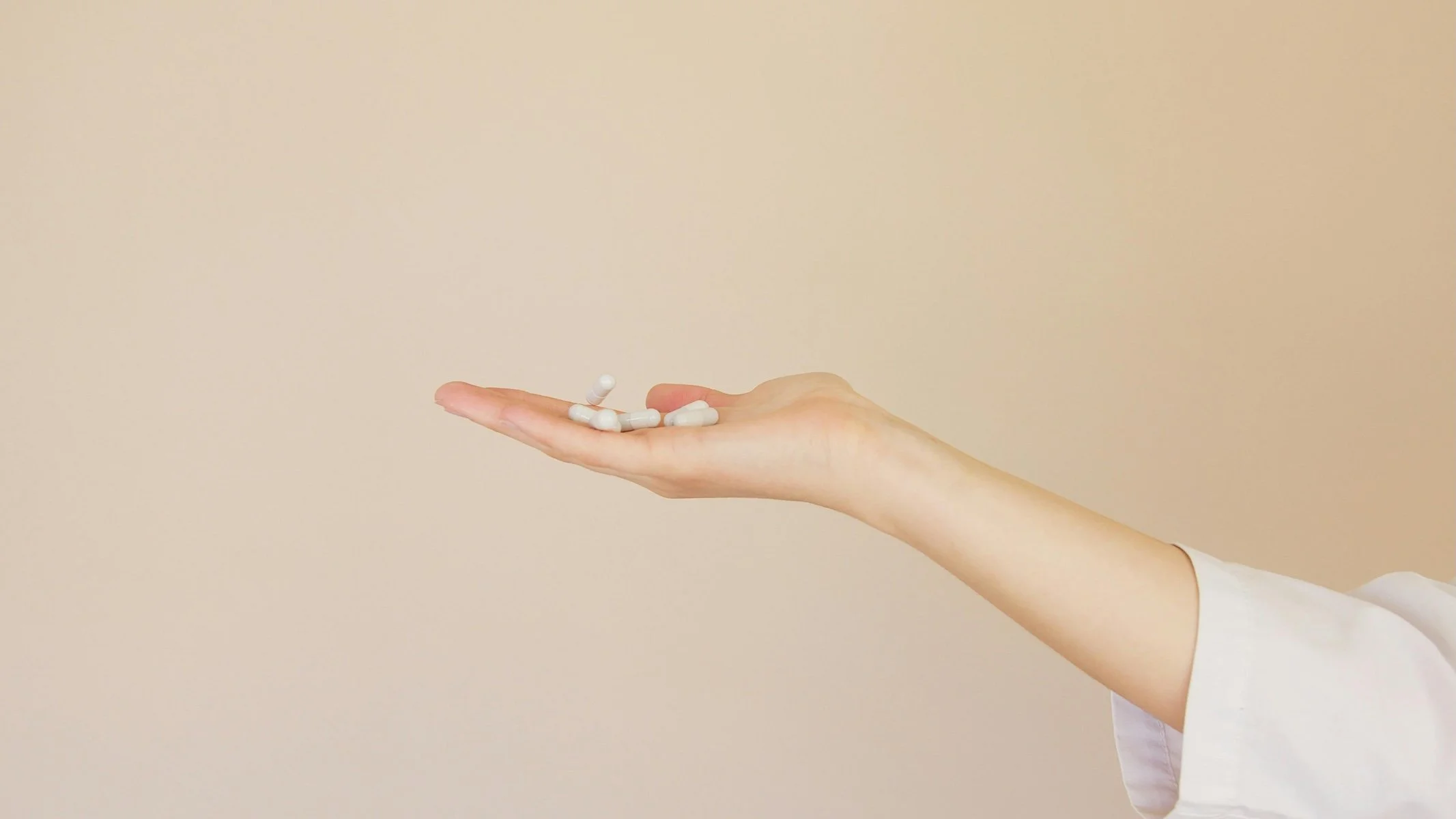 A person's hand extended sideways with pills resting on the palm against a beige background. Custom natural medicine formulations and practitioner-only supplements