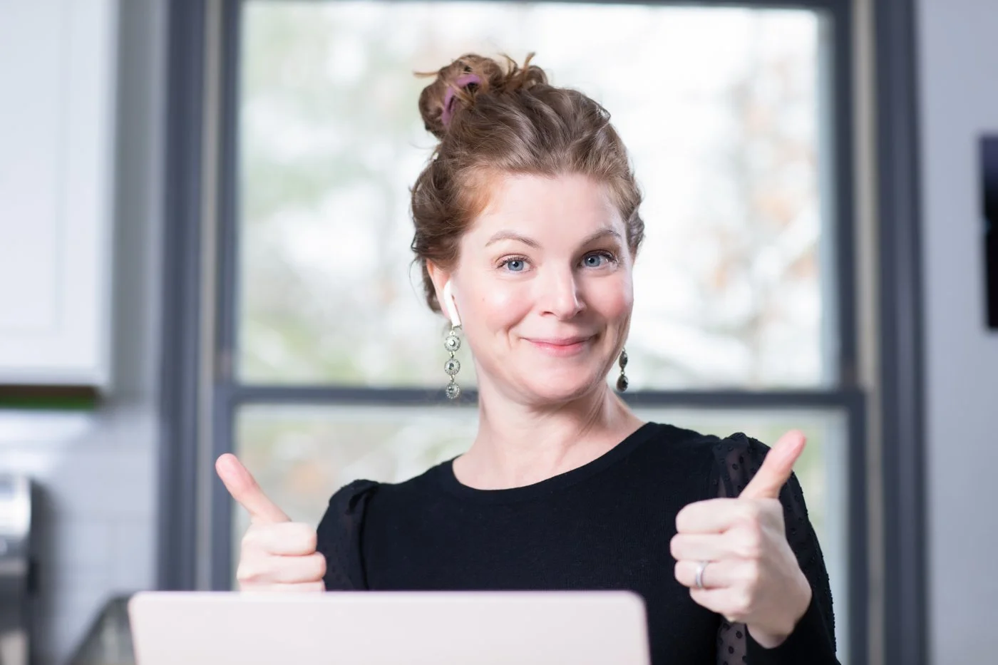 A woman with light skin and red hair in a top bun, wearing earrings and a black top, sitting in front of a laptop, smiling and giving two thumbs up.