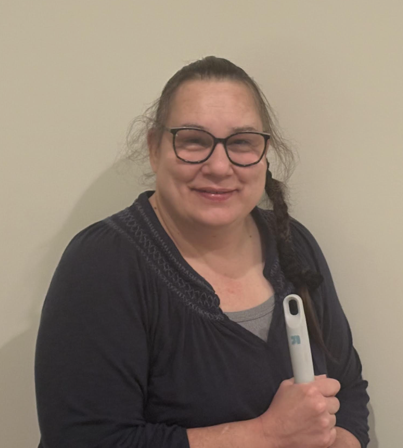 A woman with glasses and long braided hair smiling and holding a white thermometer against a plain beige wall.