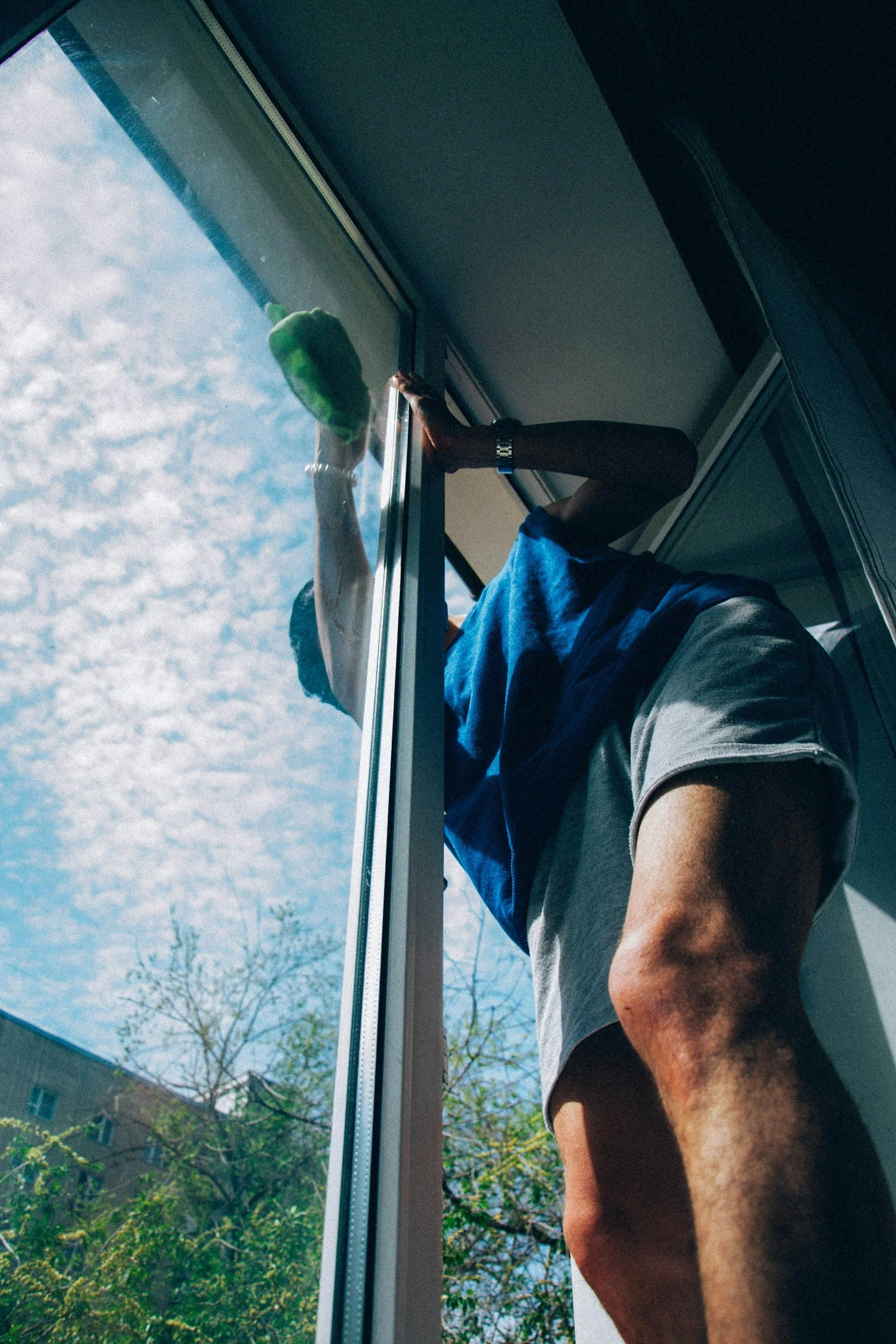 Person cleaning a window from inside using a green cleaning cloth, with a view of the blue sky and trees outside.