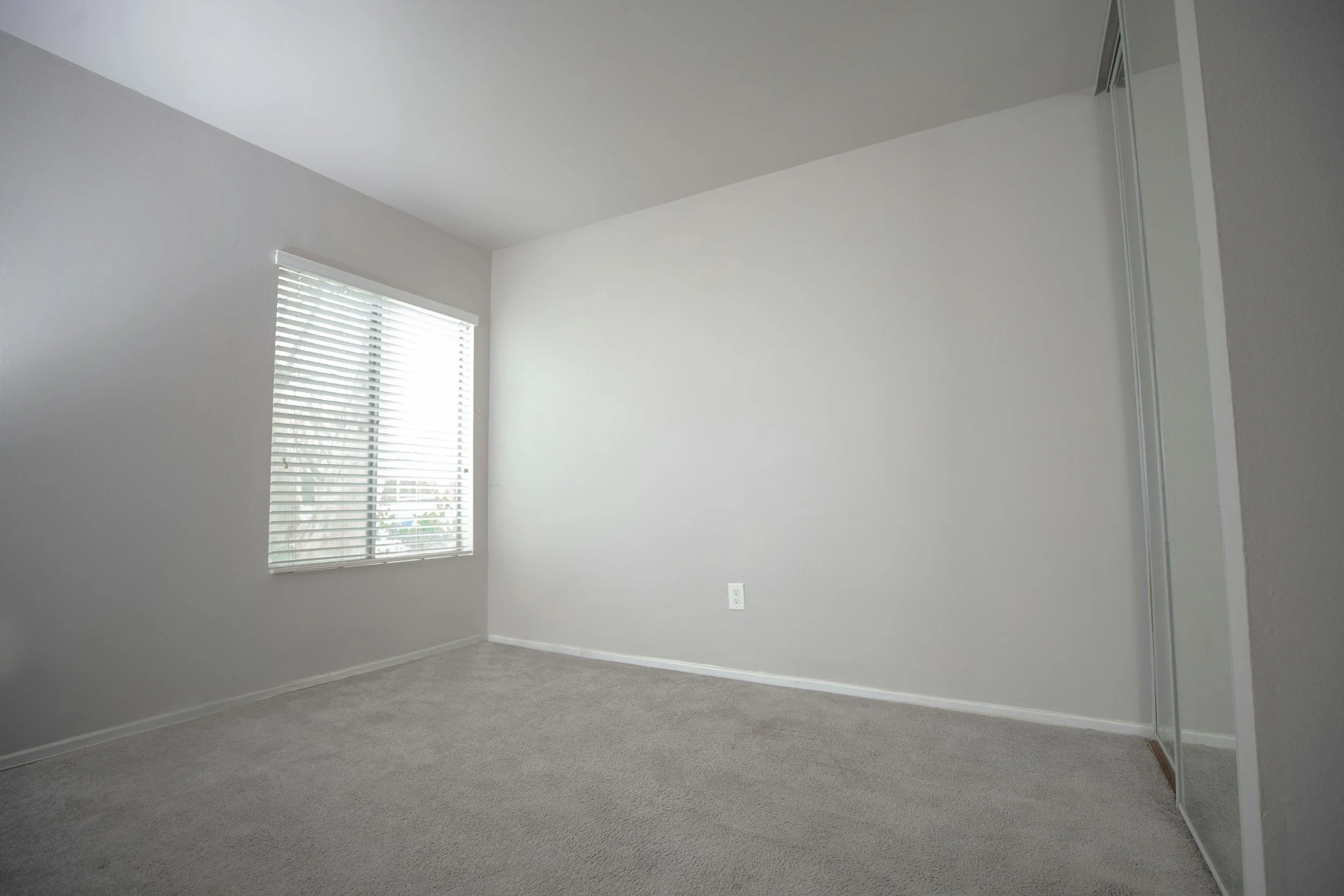Empty bedroom with light gray carpet, white walls, a window with blinds, and a mirrored closet door.