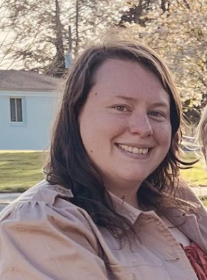 A smiling woman with shoulder-length dark brown hair outdoors, with trees and a house in the background during sunset.