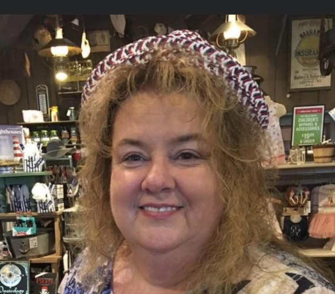 A woman with curly blond hair wearing a red, white, and blue patterned hat, smiling inside a rustic store or cafe with wooden shelves and vintage decor.