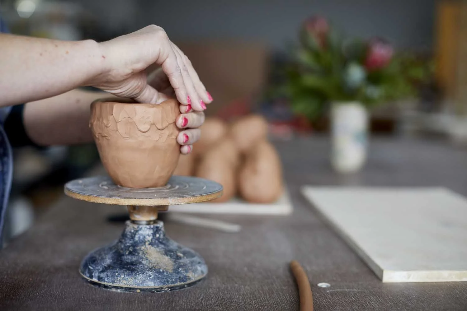 A hand built mug being scuplted by hands with pink fingernails on a banding wheel