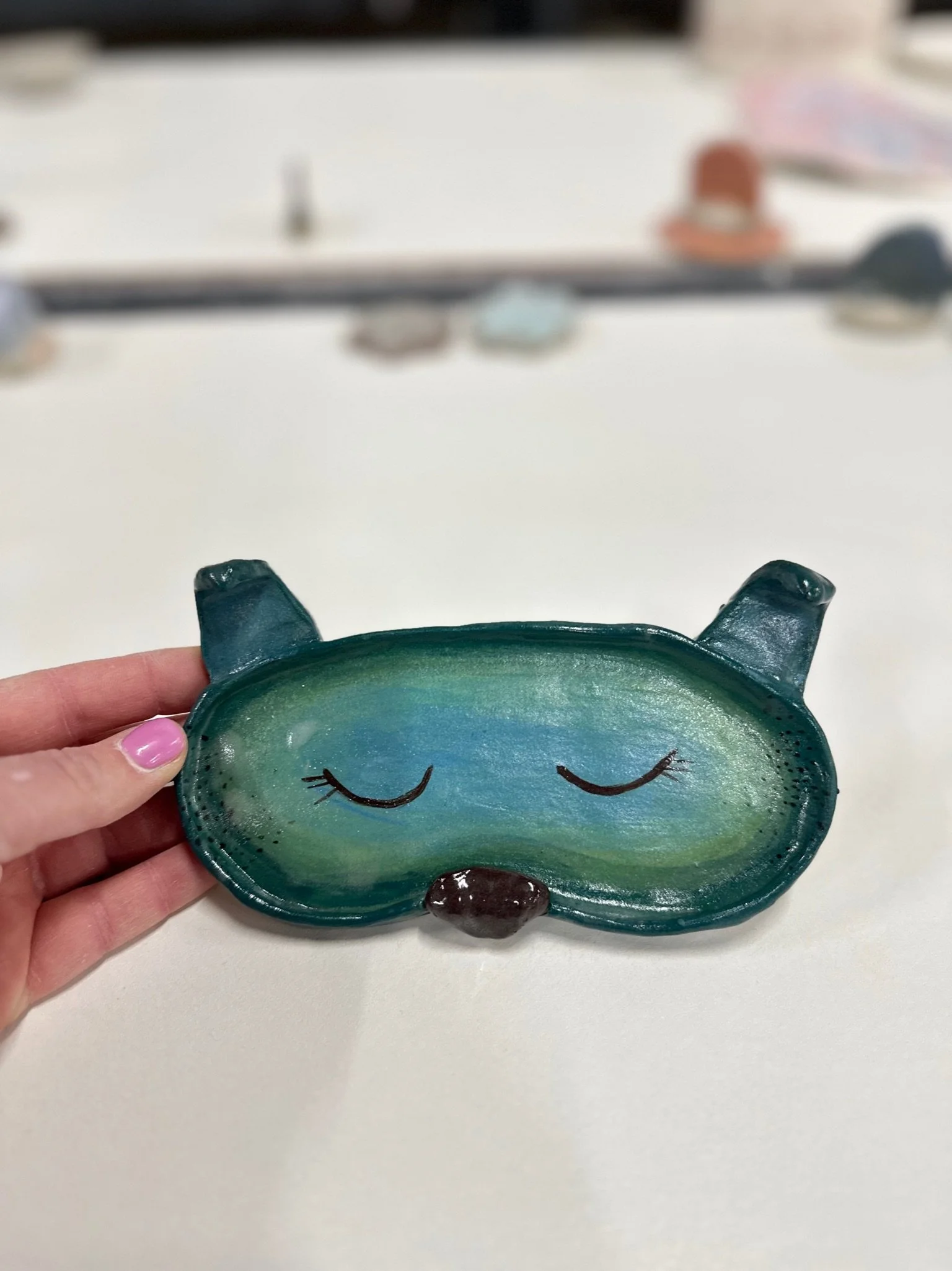 A hand holds a teal and green animal shaped glasses holder. It has folded ears, a black nose and painted on eyes and freckes