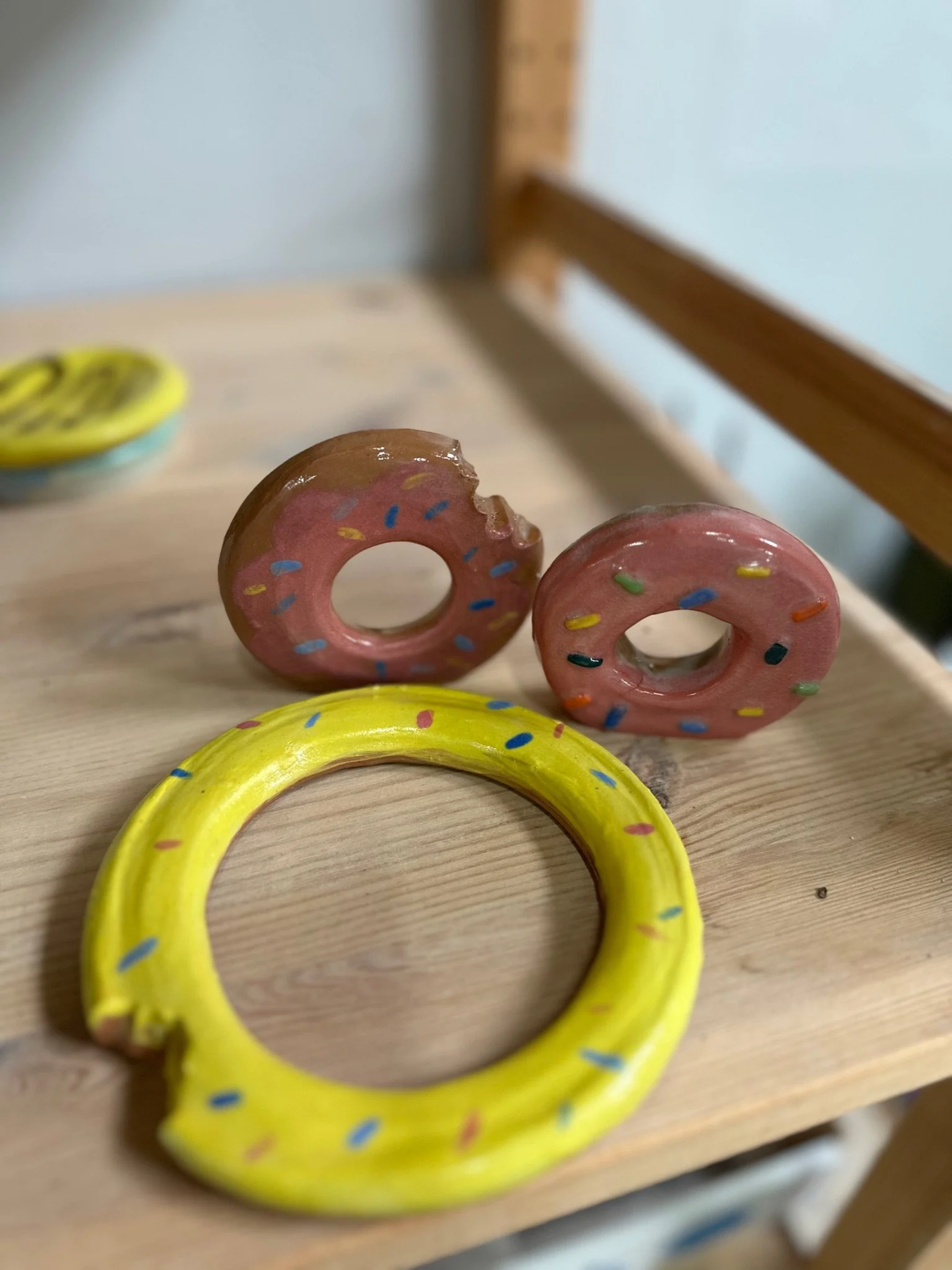 Three clay donuts on a wooden shelf. A large yellow on lays flat while two pink ones with multicoloured sprinkles stand up. One has a bite taken out of it.