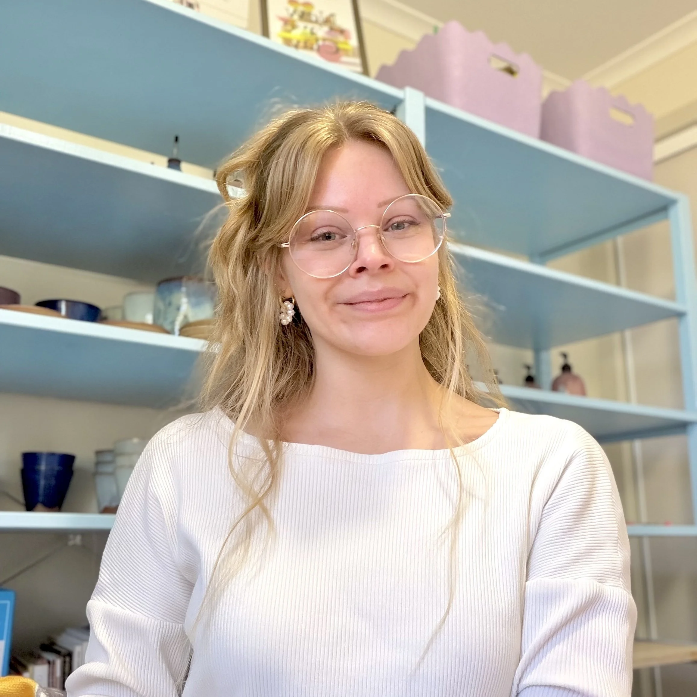 A portrait of a blonde hair woman with round silver rimmed glasses, with a closed mouth smile. In the background are pale blue shelves