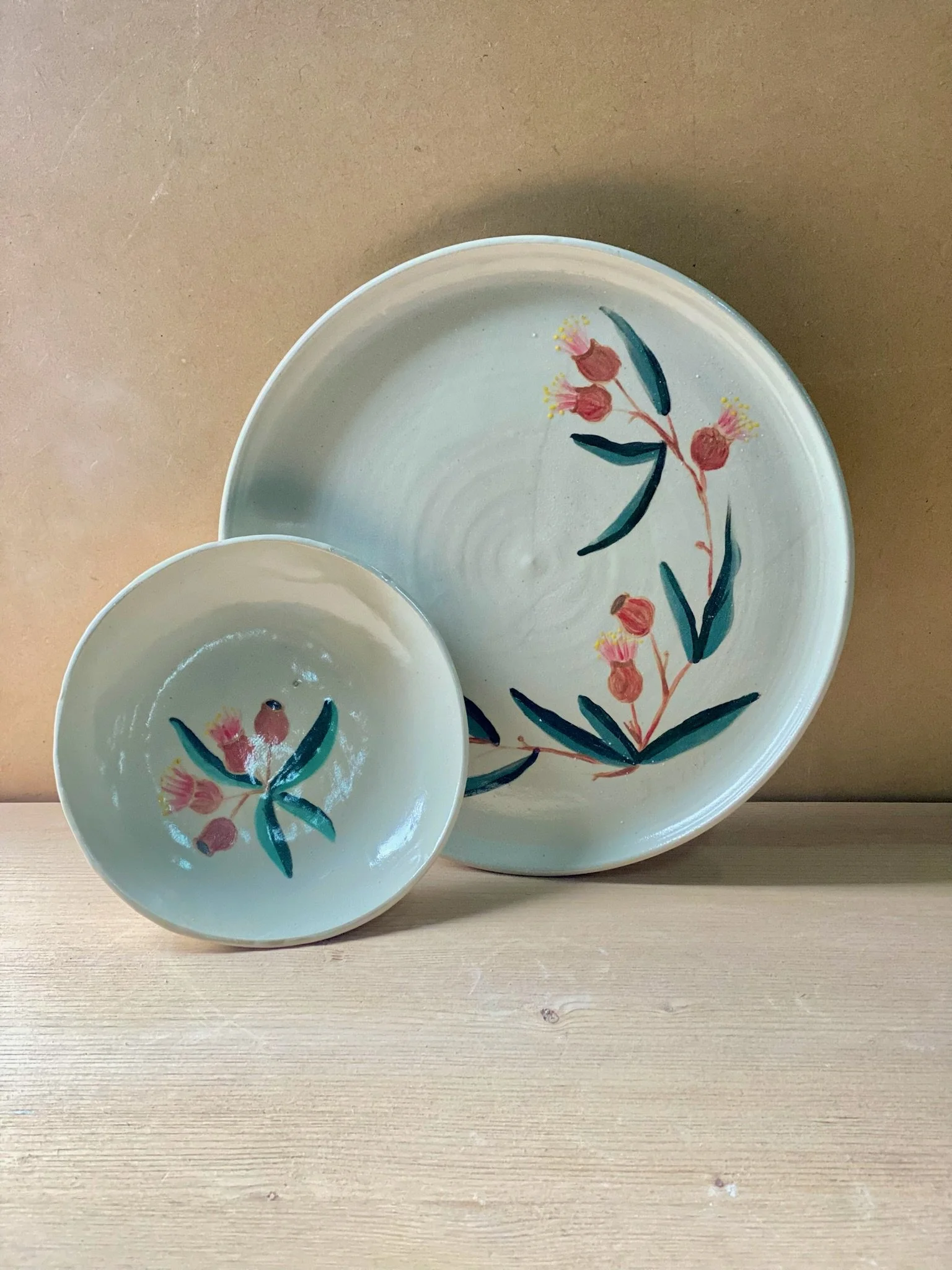 A large plate in the background and a bowl in the foreground set on a wood backdrop. Both pieces are painted with green eucalyptus leaves and red, pink and yellow gum flowers.