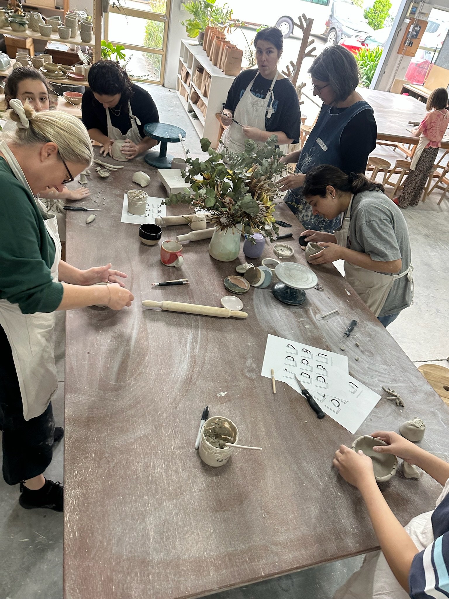 A group of people wearing aprons standing around the table, hand building with clay. The table has sample pieces, glaze samples, tools and a vase with flowers on it