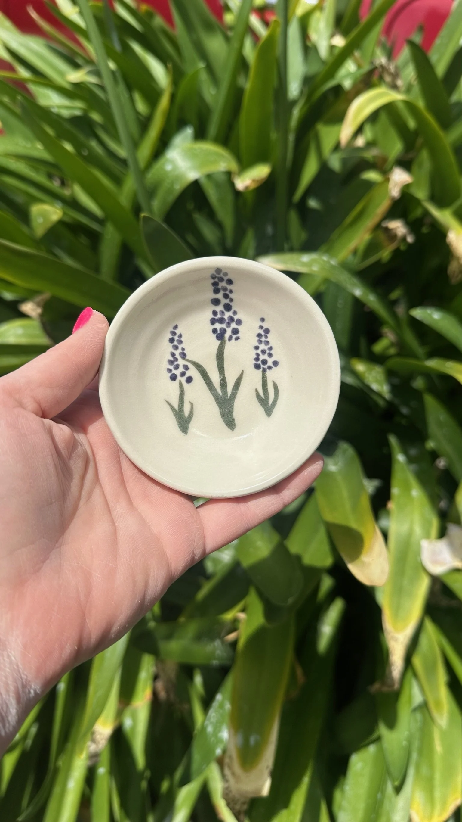 A hand holds a small beige trinket dish with purple vertical flowers with green stems painted on