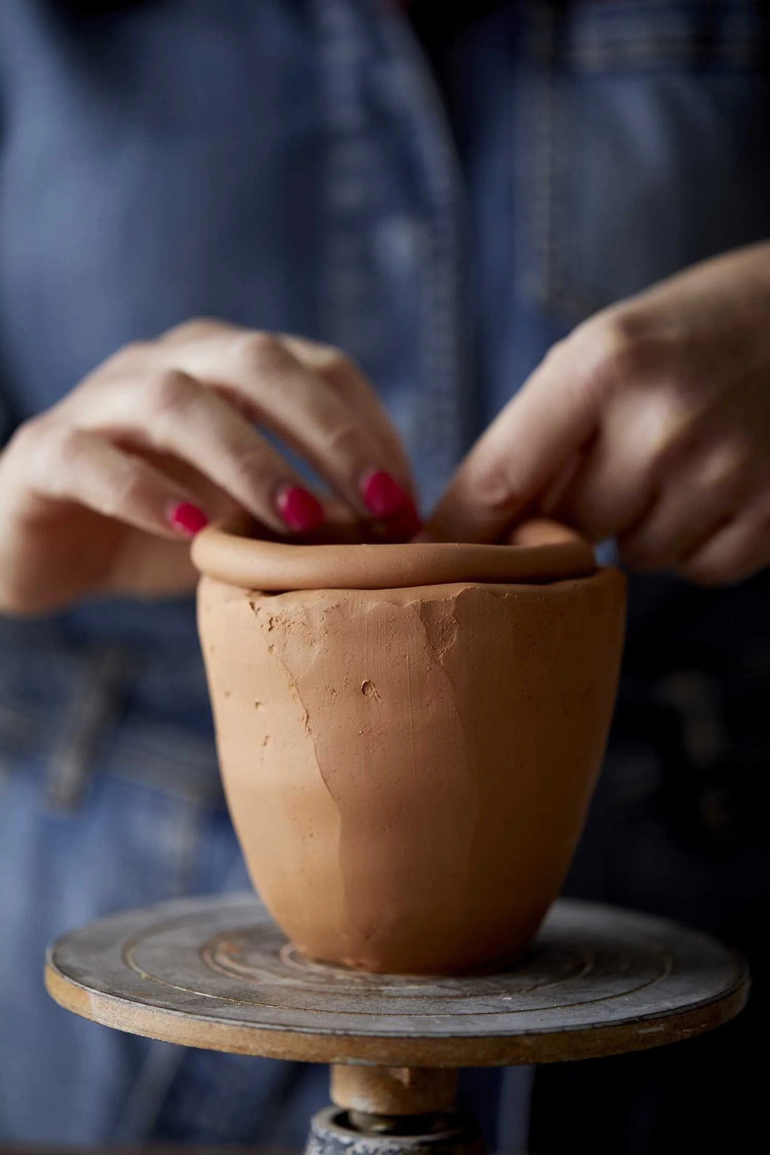 An image of a handbuilt mug in a clau colour with a coil on top. A pair of hands is blurred in the background with pink fingernails, blending the coil in