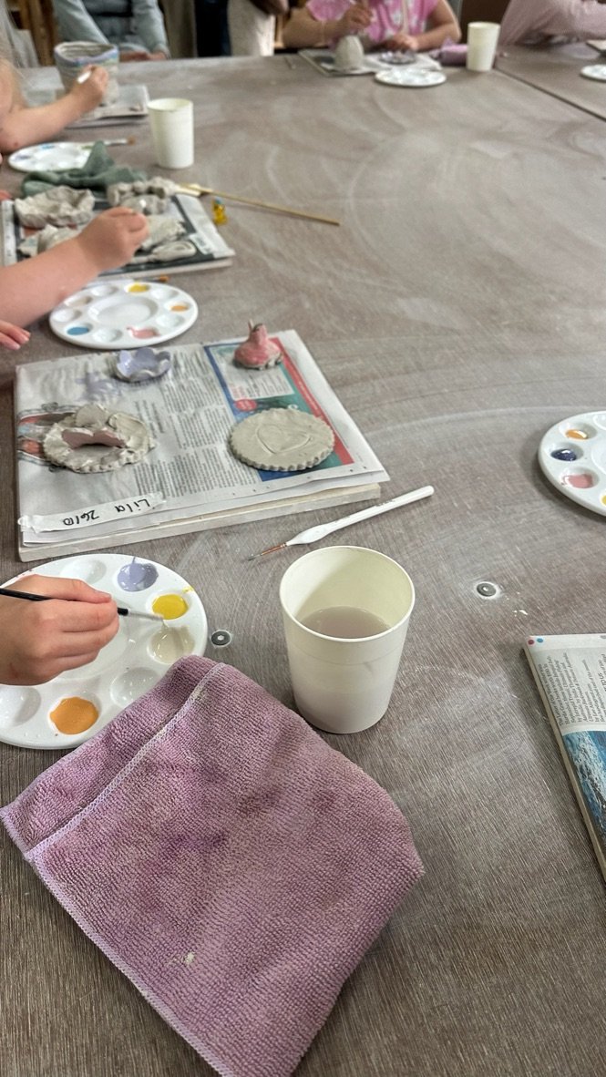 A table with paint pallettes, and clay creations on boards, you can see small hands painting