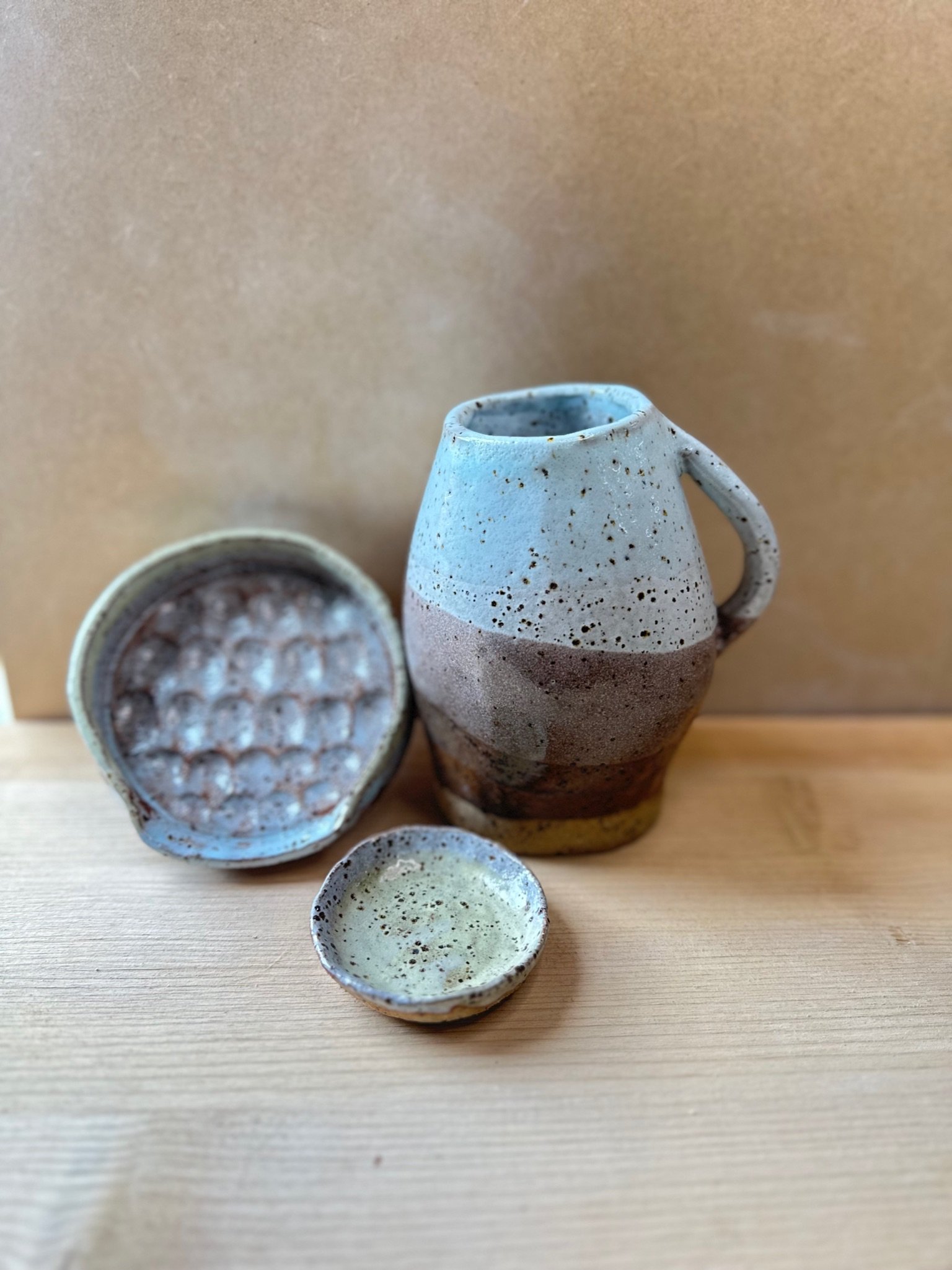 A large jug in pale blue and brown is in the foreground with a small green-blue round trinket dish. In the blurred background sits a textured spoon rest