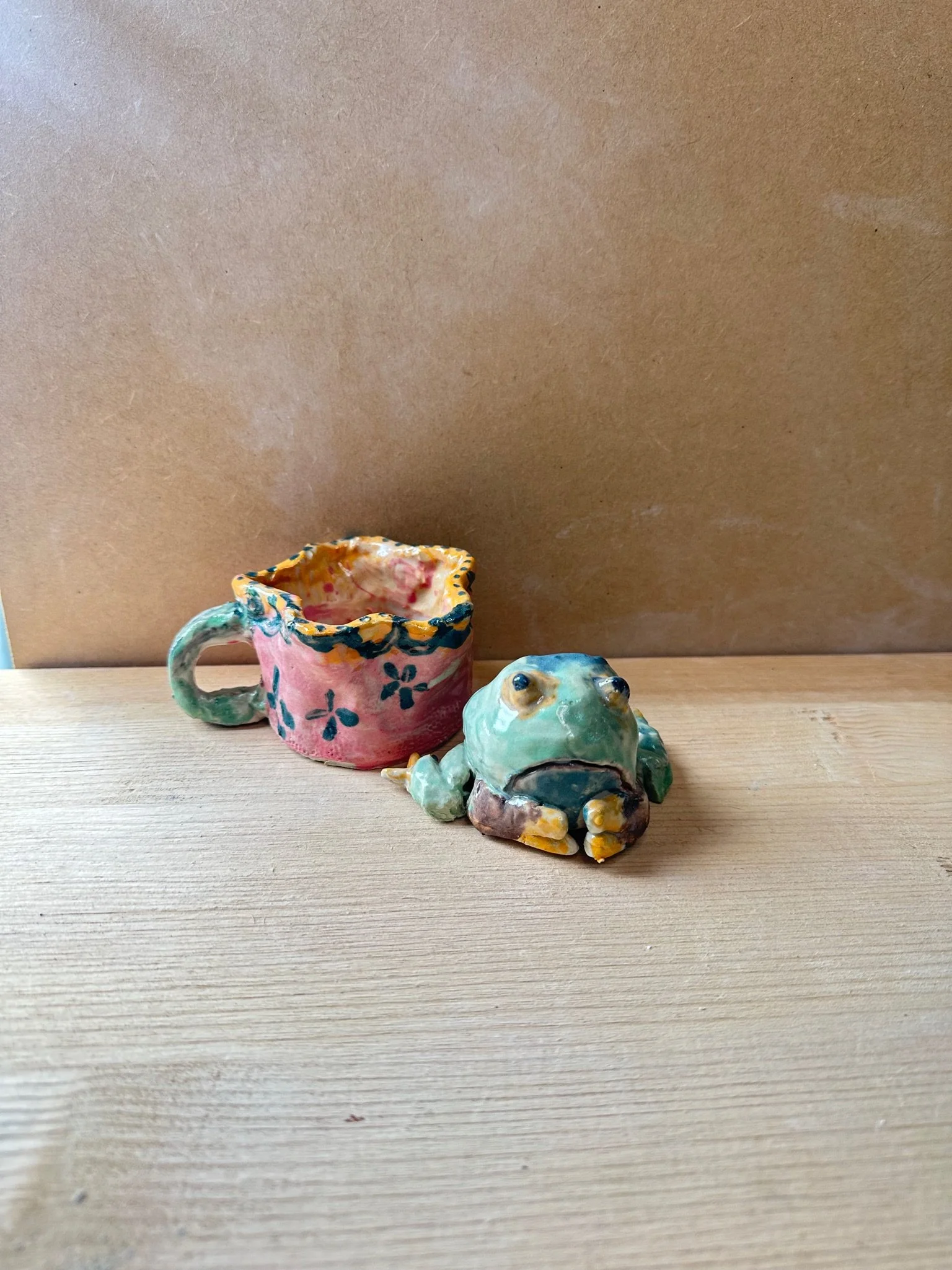 Two pottery pieces on a wooden table. A brightly coloured mug with green handles and blue flowers on a pink background with an orange lip. In the foreground is a green frog with brown and yellow legs and blue buggy eyes.