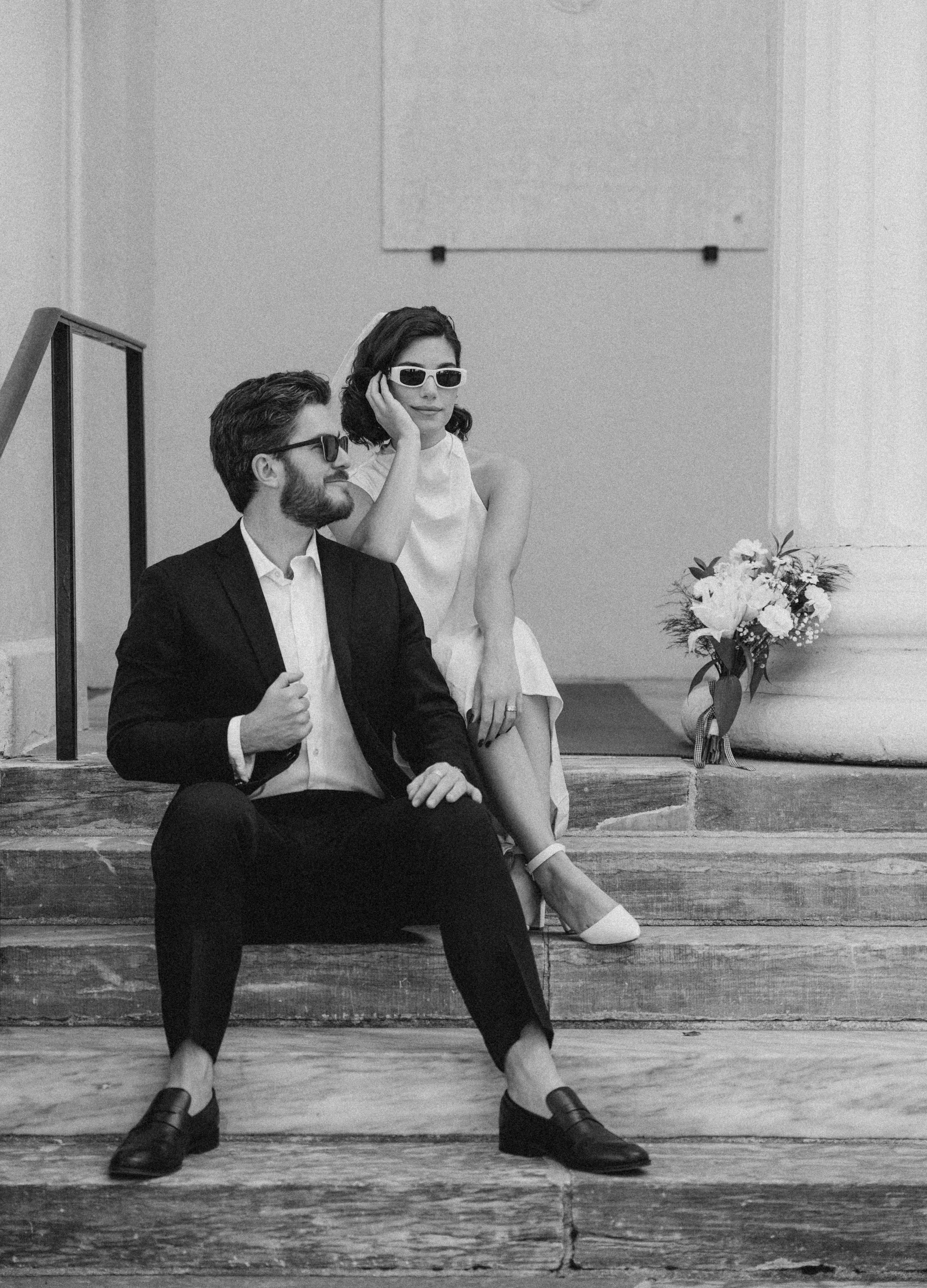 A black-and-white photo of a man and woman sitting on stone steps, both wearing sunglasses. The woman is resting her head on her hand, and the man is adjusting his sleeve. There is a bouquet of flowers nearby.