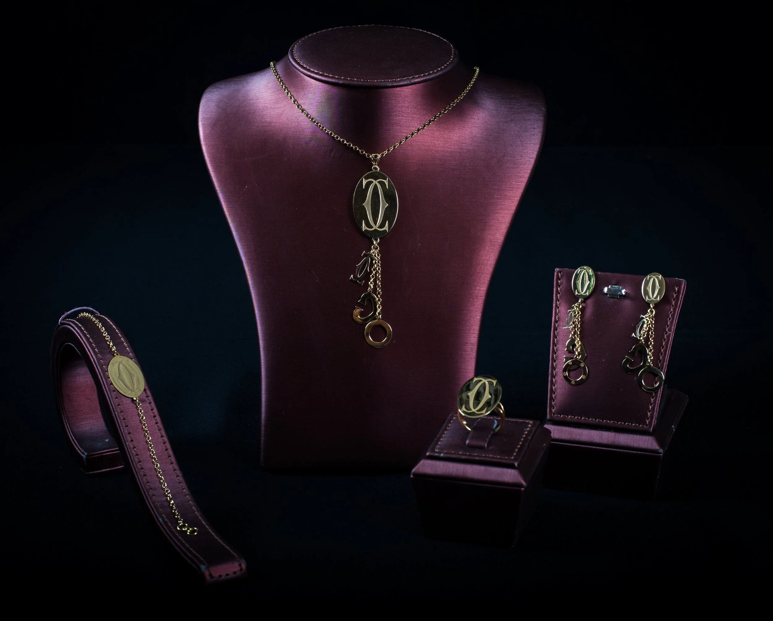 Gold jewelry set including a necklace, earrings, a ring, and a bracelet, displayed on burgundy jewelry stands and a burgundy box for the ring, against a black background.