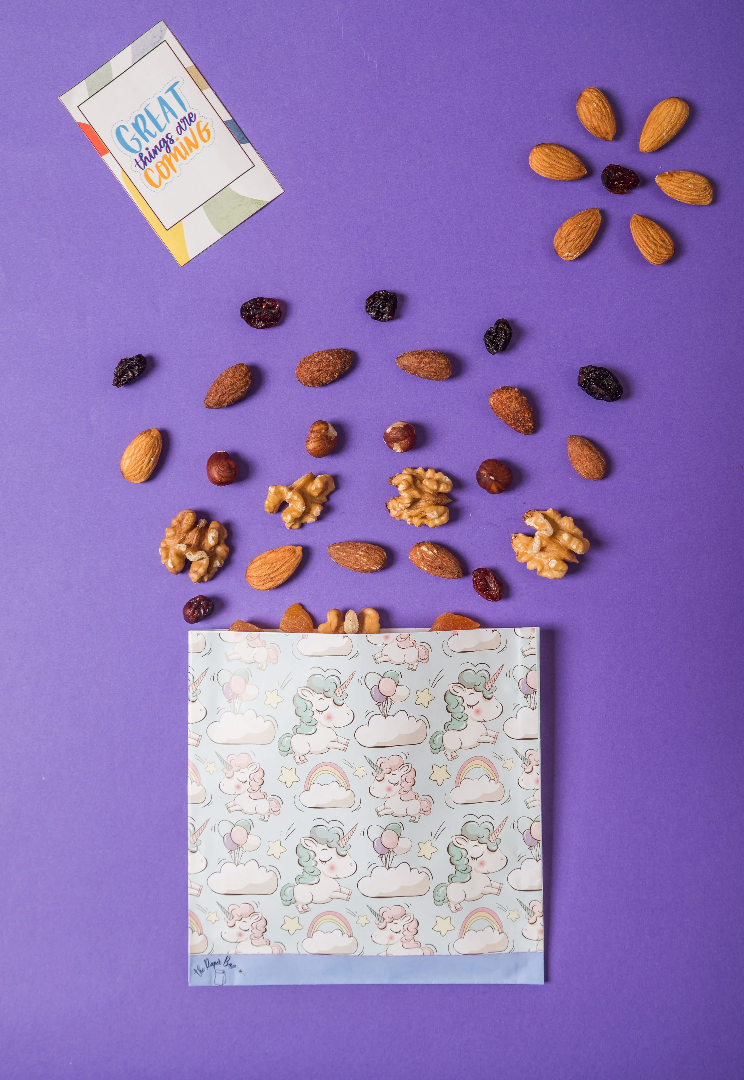 A collection of nuts, dried fruits, and a unicorn-themed paper bag with a purple background, along with a card that reads "Great things are coming" placed in the top left corner.