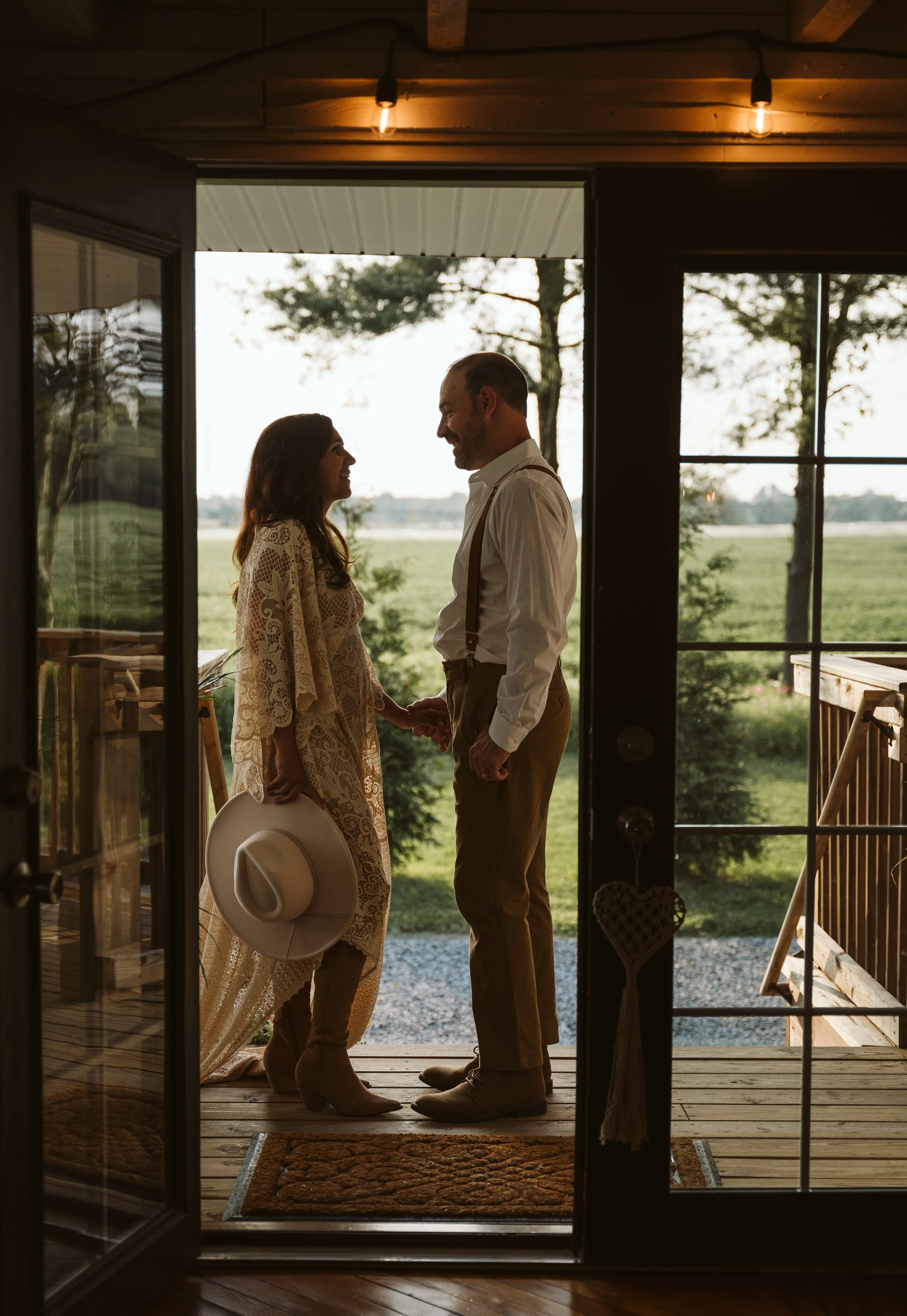 A couple holding hands and smiling at each other on a porch, with the woman holding a hat, seen through an open door leading outside, with trees and fields in the background during daytime.