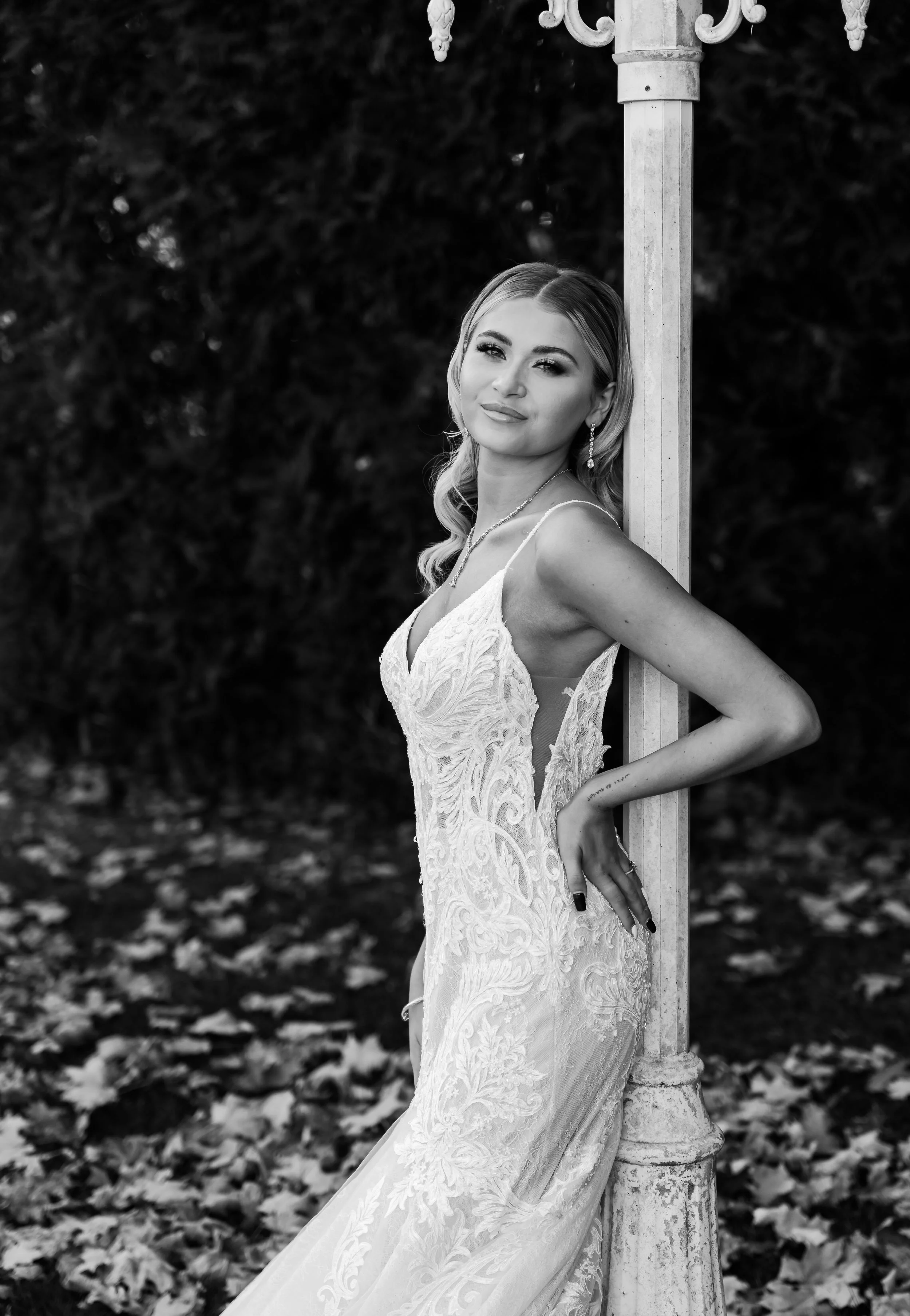 A woman in an elegant lace dress with spaghetti straps, standing outdoors next to a decorative streetlamp post, with fallen leaves on the ground and dark foliage in the background.