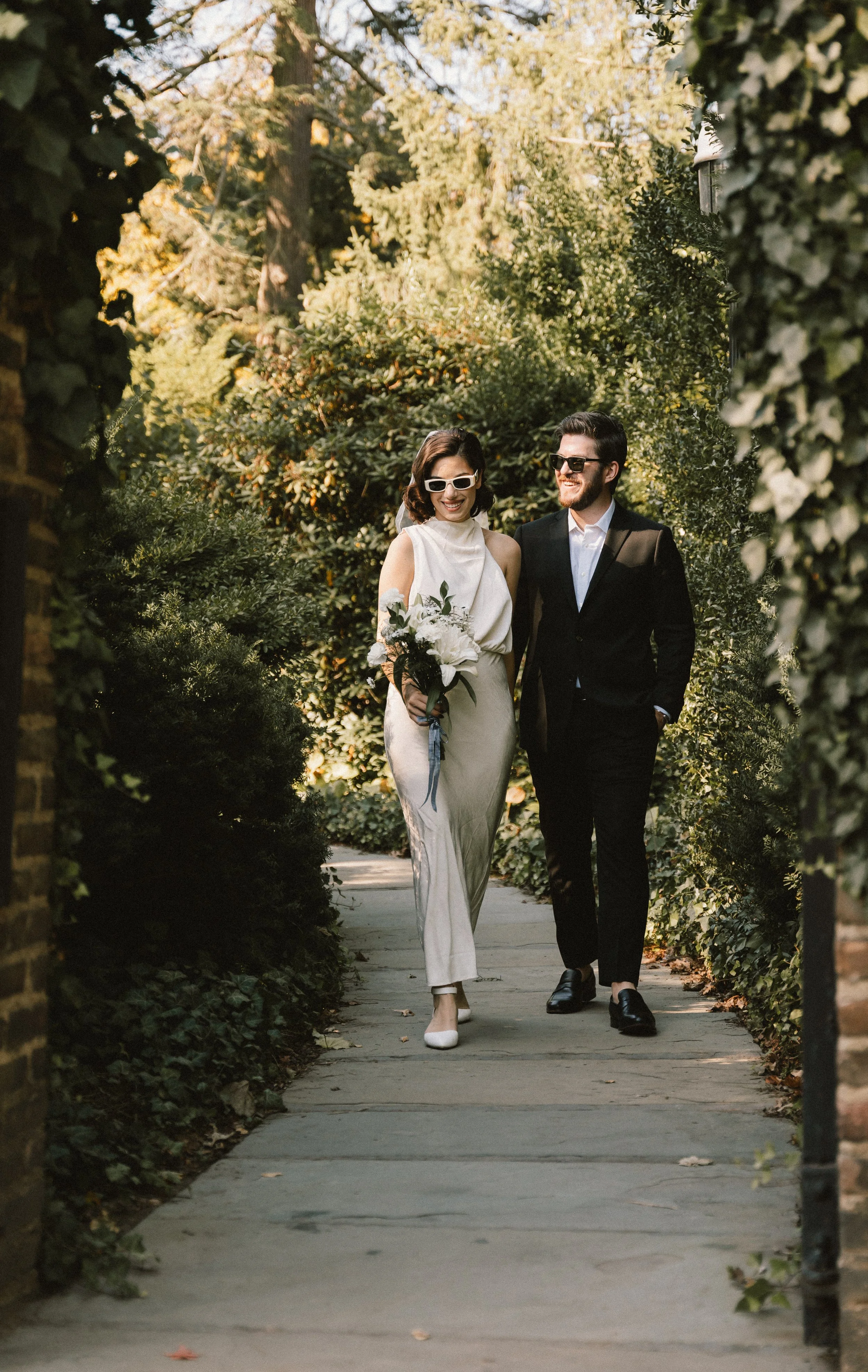 A woman in a cream-colored dress holding a bouquet walking with a man in a black suit on a garden pathway, both wearing sunglasses and smiling.