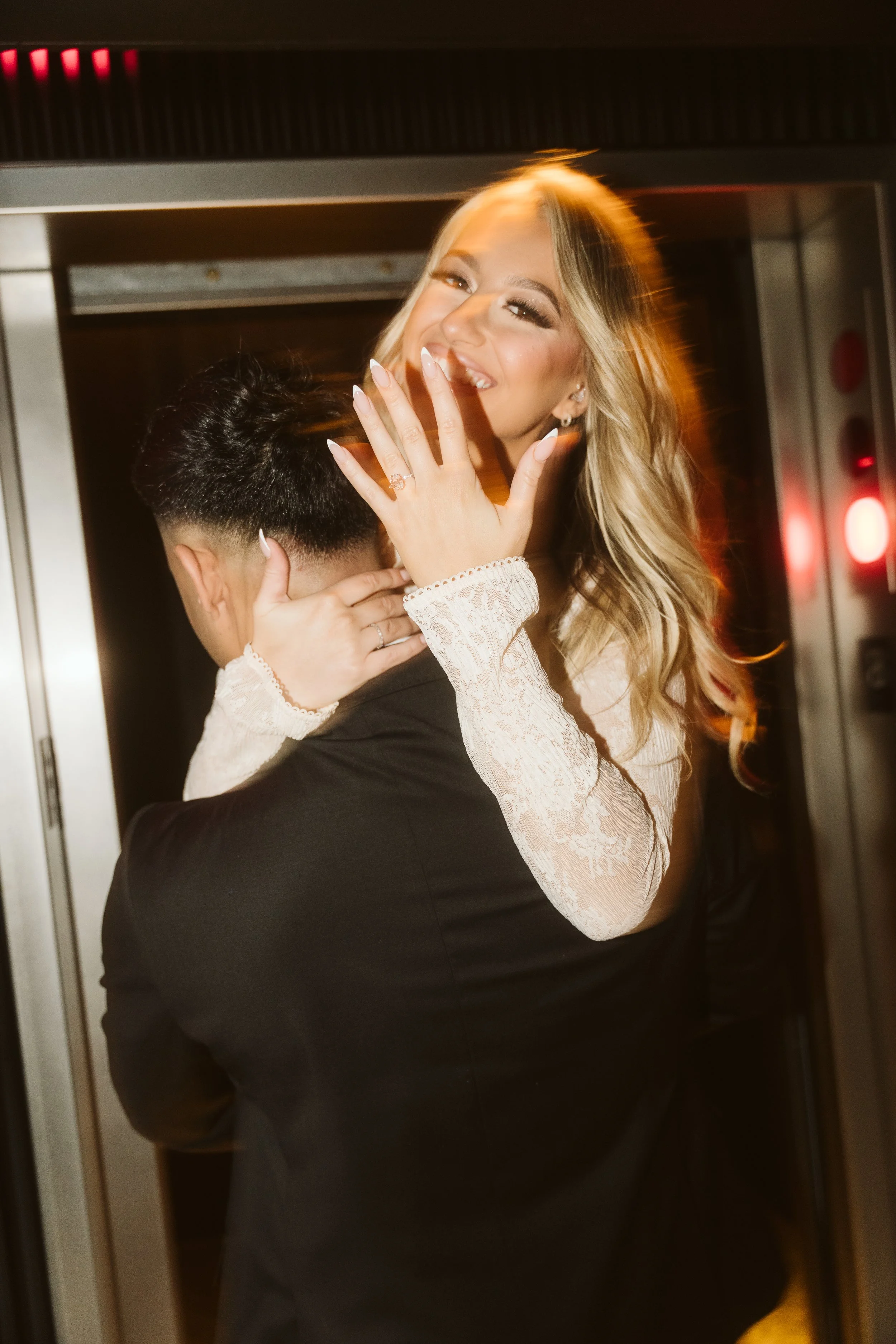 A woman with long blonde hair, wearing a white lace dress, being lifted by a man in a black suit inside an elevator. She is smiling and covering her mouth with her hand, showing a ring on her finger.