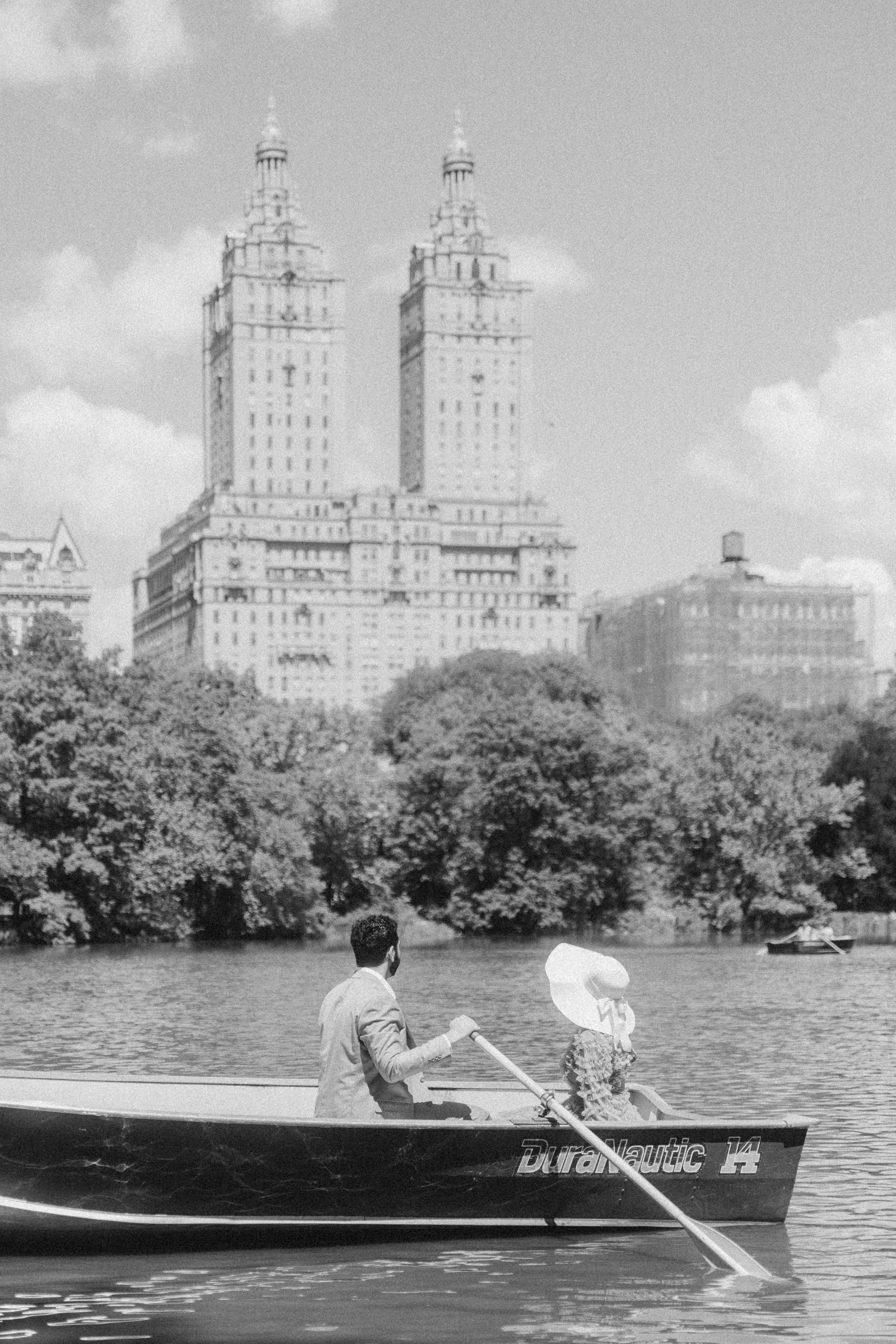 A man and a woman sitting in a boat on a river with tall buildings and trees in the background.