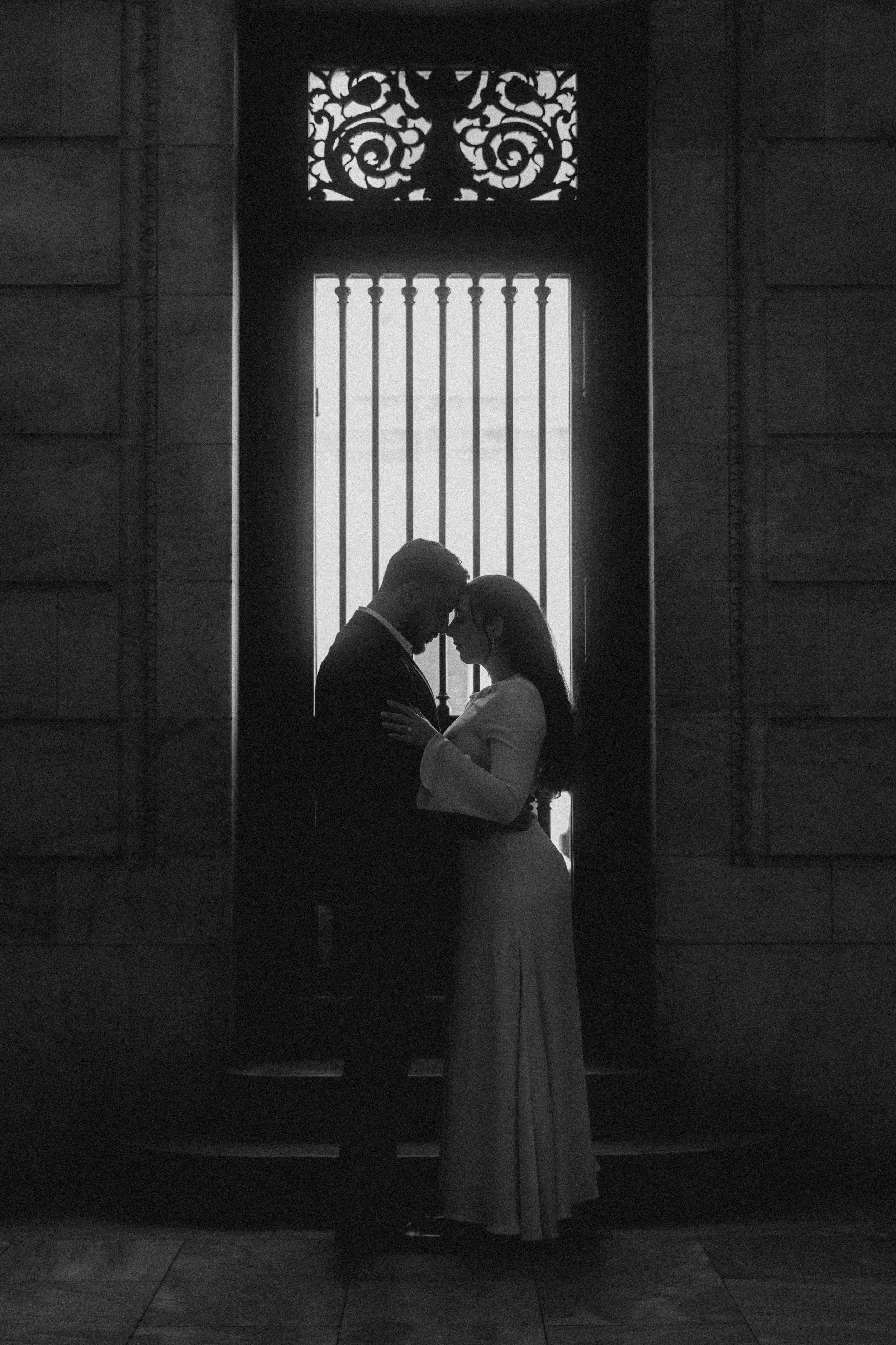 Silhouetted couple standing close, touching foreheads and holding each other inside a building with decorative iron window grilles in the background.