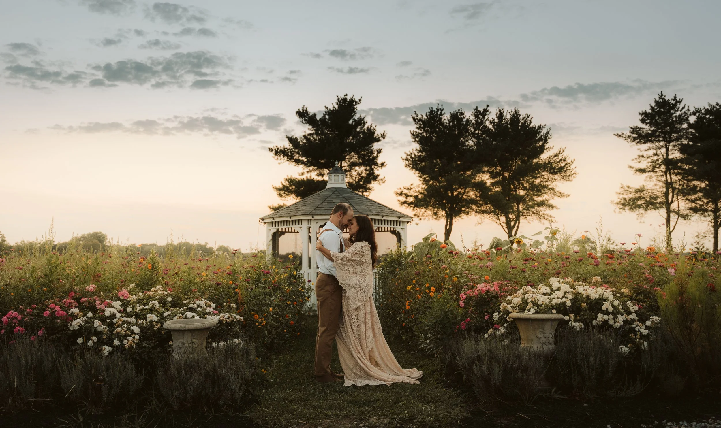 A couple standing close in a garden at sunset, surrounded by flowers and trees, with a gazebo in the background.