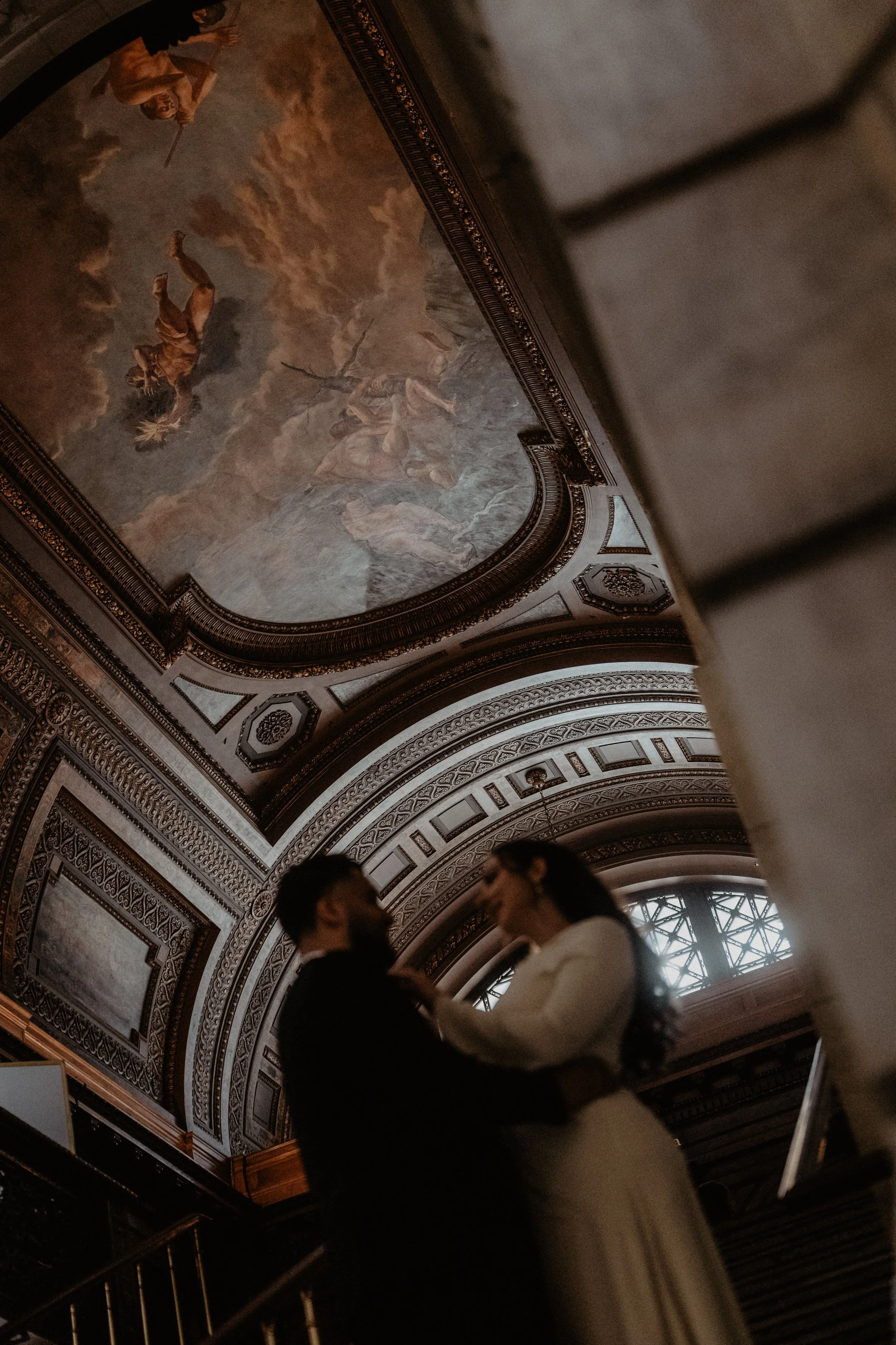 A blurred silhouette of a couple dancing in a grand, ornately decorated hall with a detailed painted ceiling and large window.