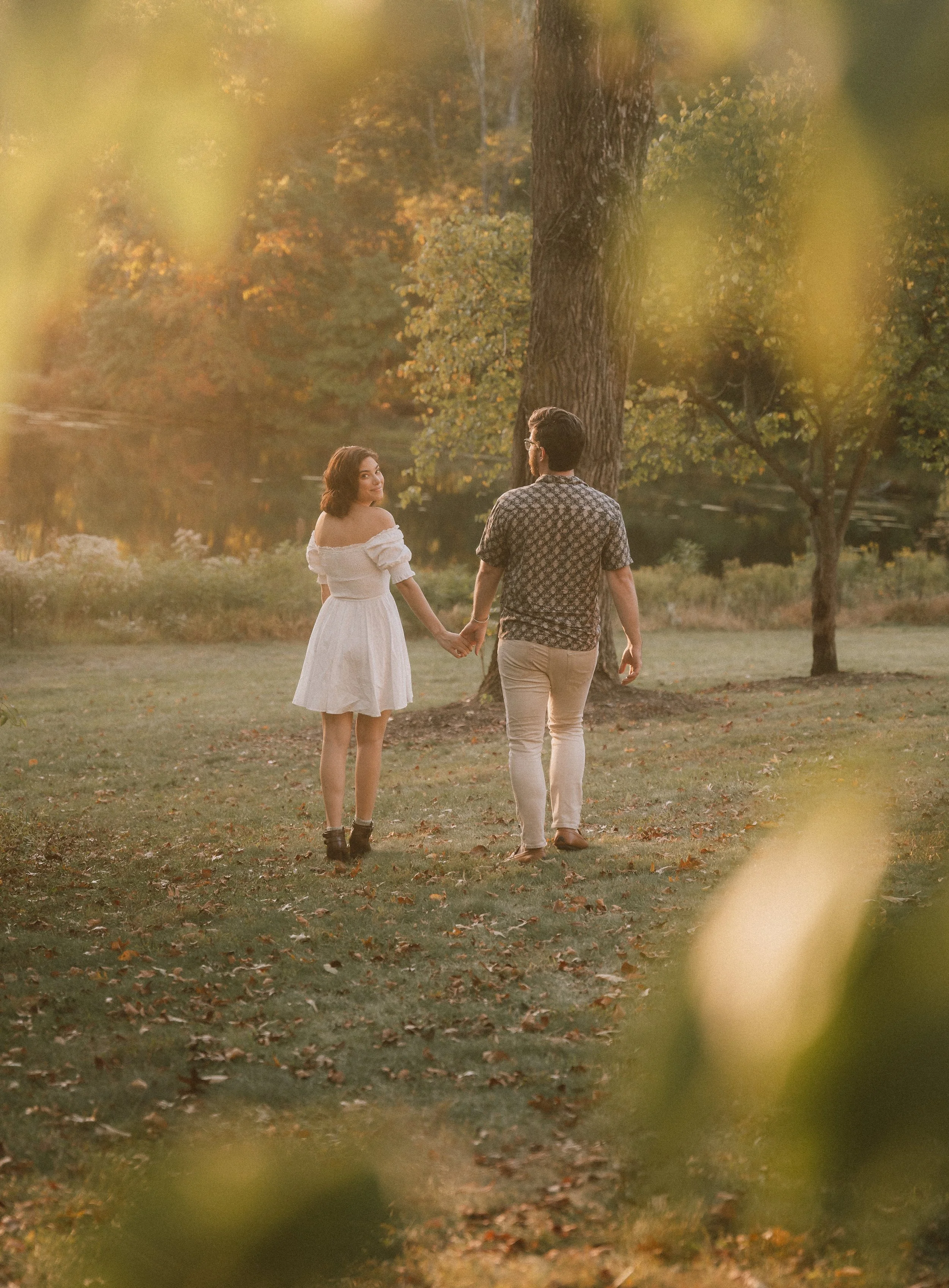 A couple holding hands walking in a park during sunset. The woman is wearing a white dress and boots, and the man is wearing a patterned shirt and light-colored pants. There are trees with autumn leaves around them.