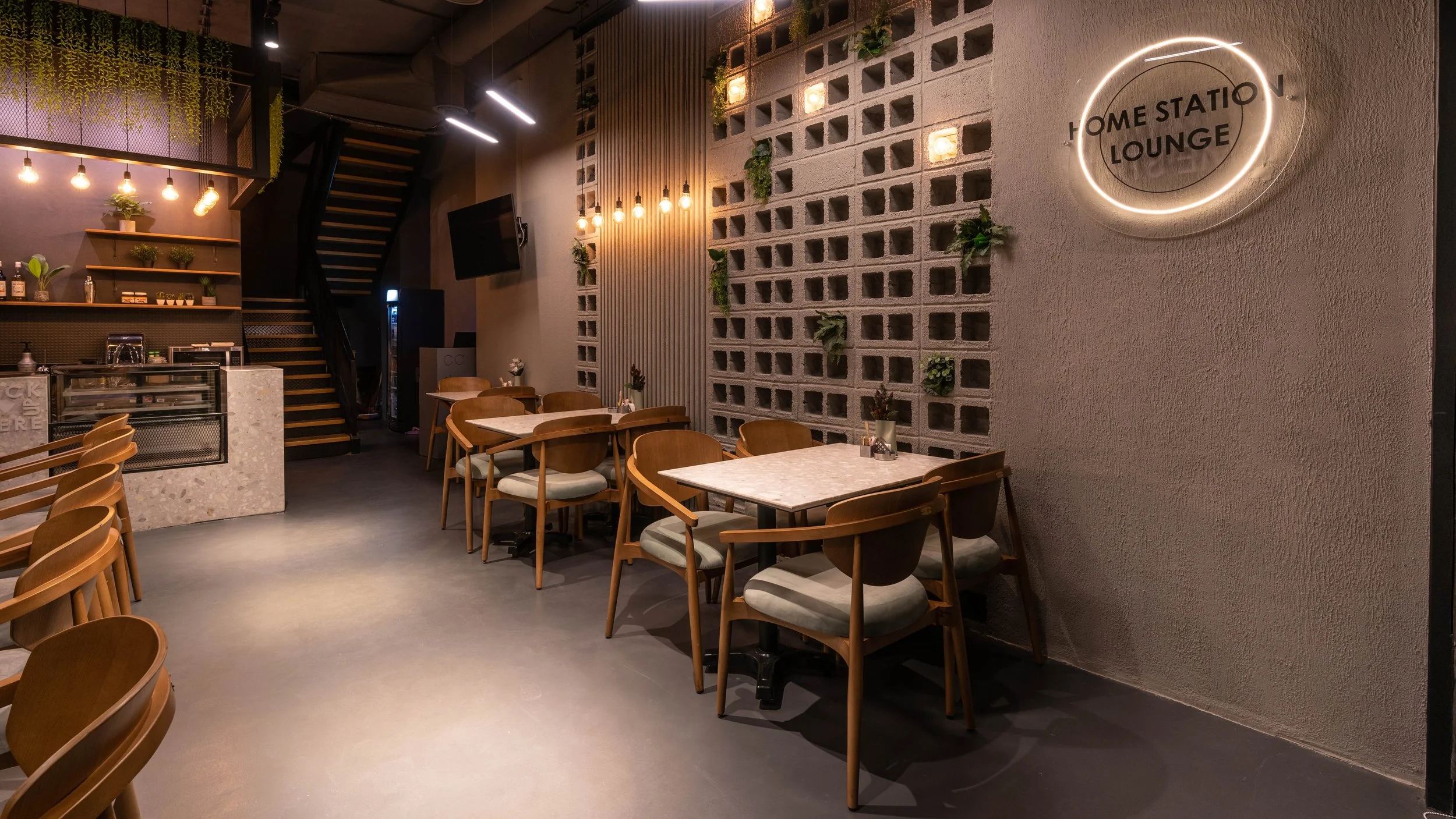 Interior of a modern, cozy lounge with wooden chairs and marble-top tables, decorative wall with plants, warm lighting, and a neon sign reading 'Home Station Lounge'.