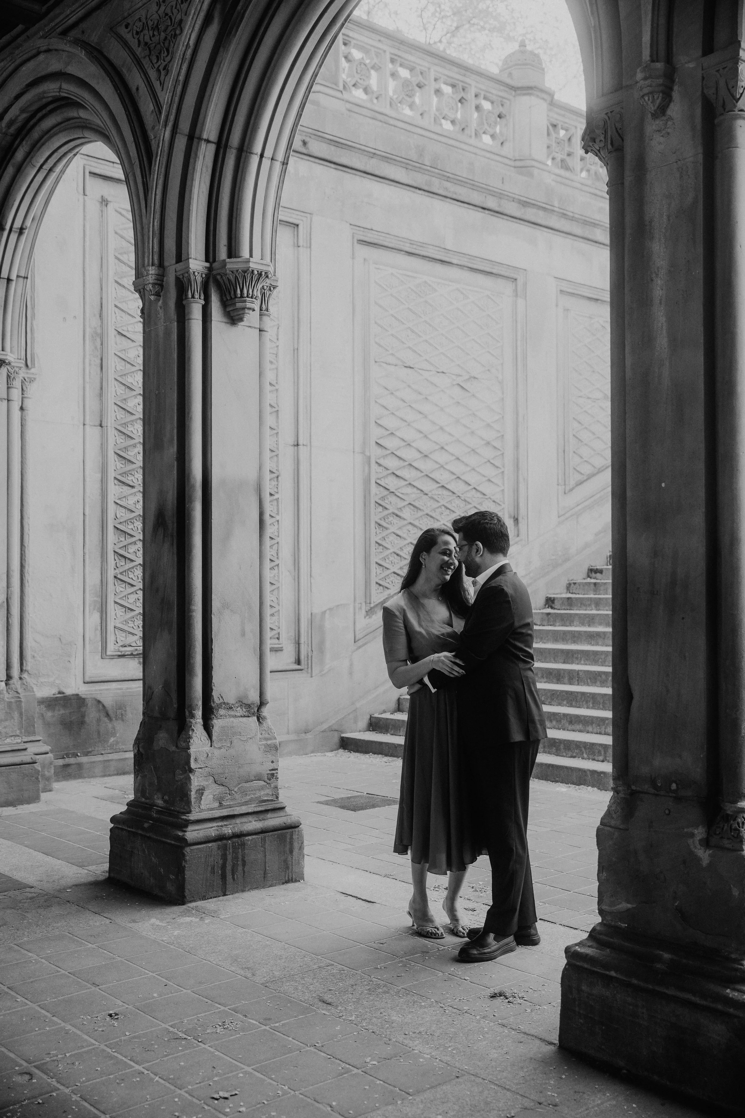 A black-and-white photo of a couple smiling and holding each other under a stone archway with detailed columns and stairs in the background.