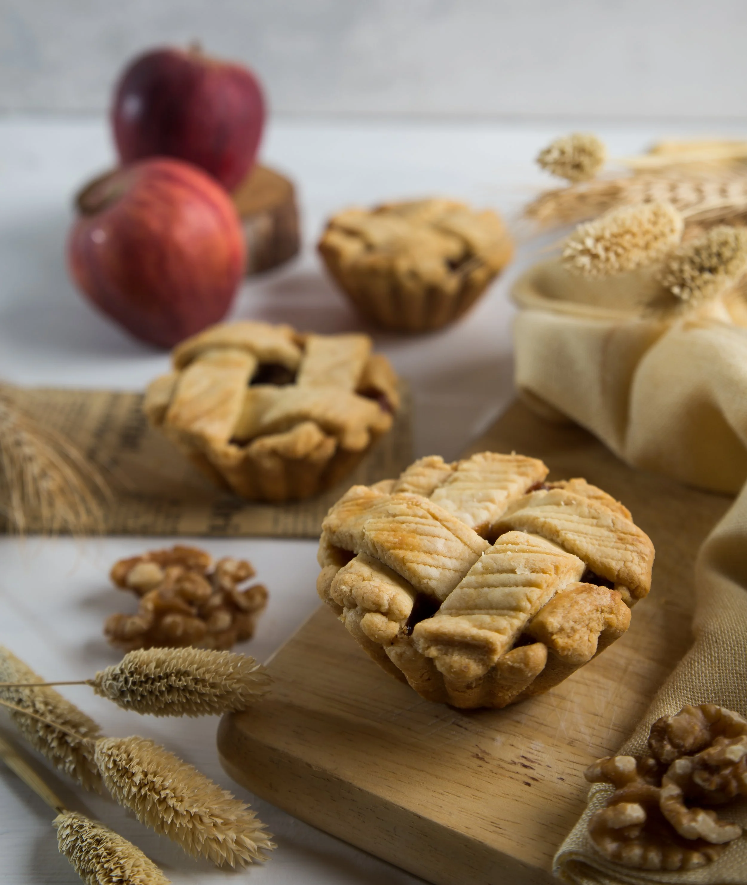Close-up of homemade apple crisp pies with a picnic table of apples, walnuts, and dried wheat stalks in the background.