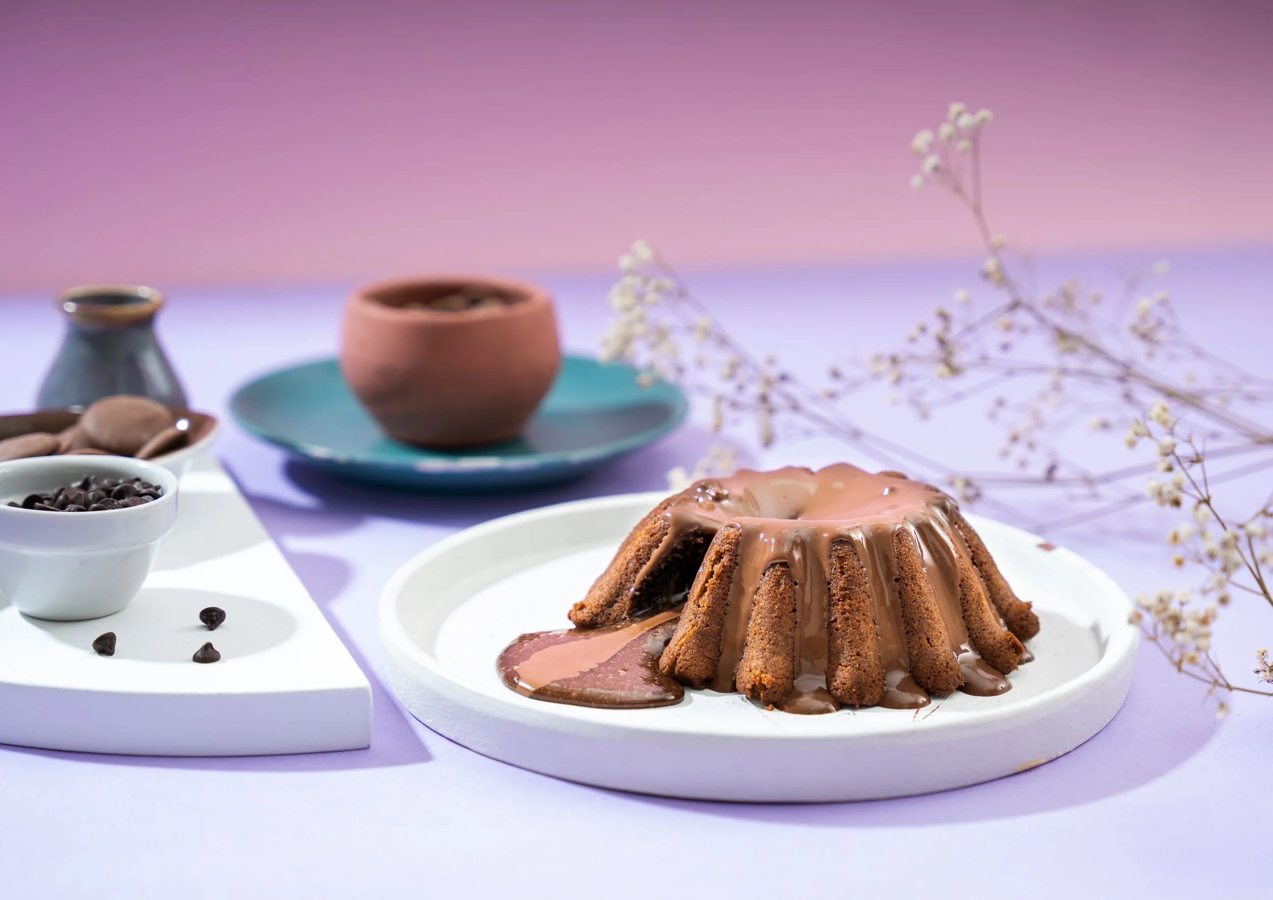 Chocolate glazed bundt cake on a white plate, with chocolate sauce dripping down the sides, surrounded by small bowls of chocolate chips and coffee beans, with decorative flowers in the background.