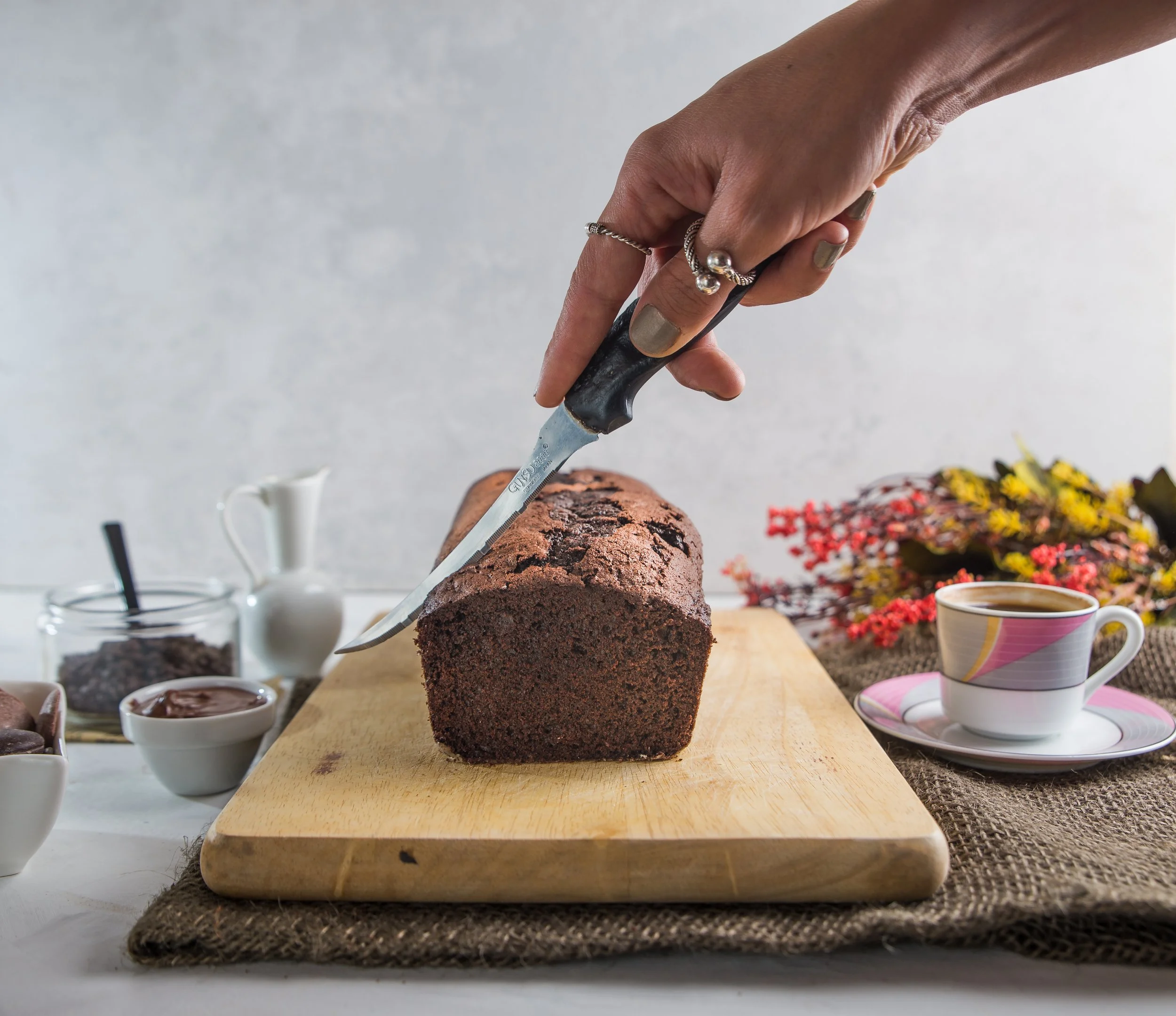 A hand with rings is slicing a chocolate loaf cake on a wooden cutting board, with a cup of coffee, bowls of chocolate, and flowers in the background.