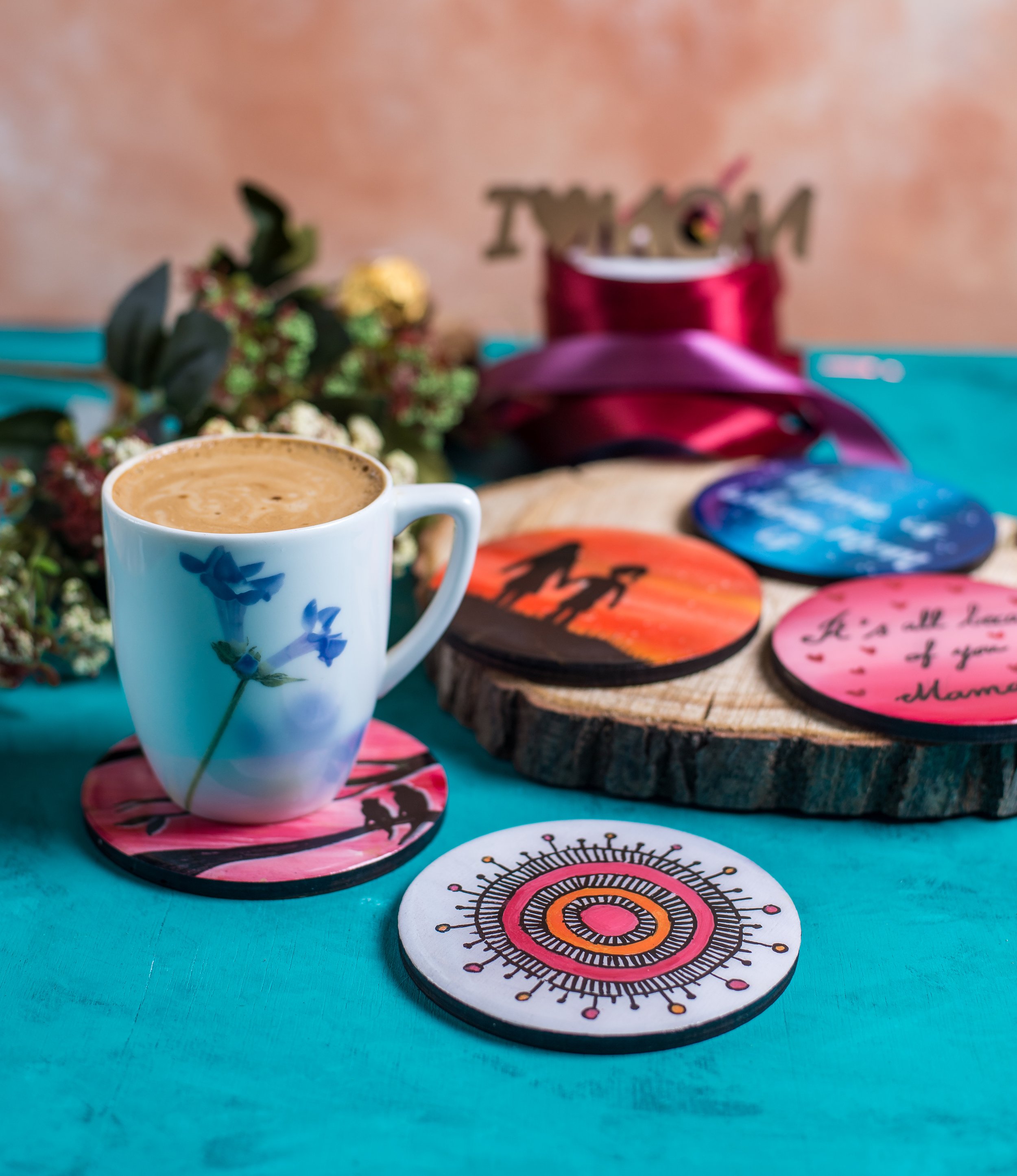 A coffee mug with blue flower design on a pink coaster, on a teal surface with colorful coasters and a floral arrangement in the background.