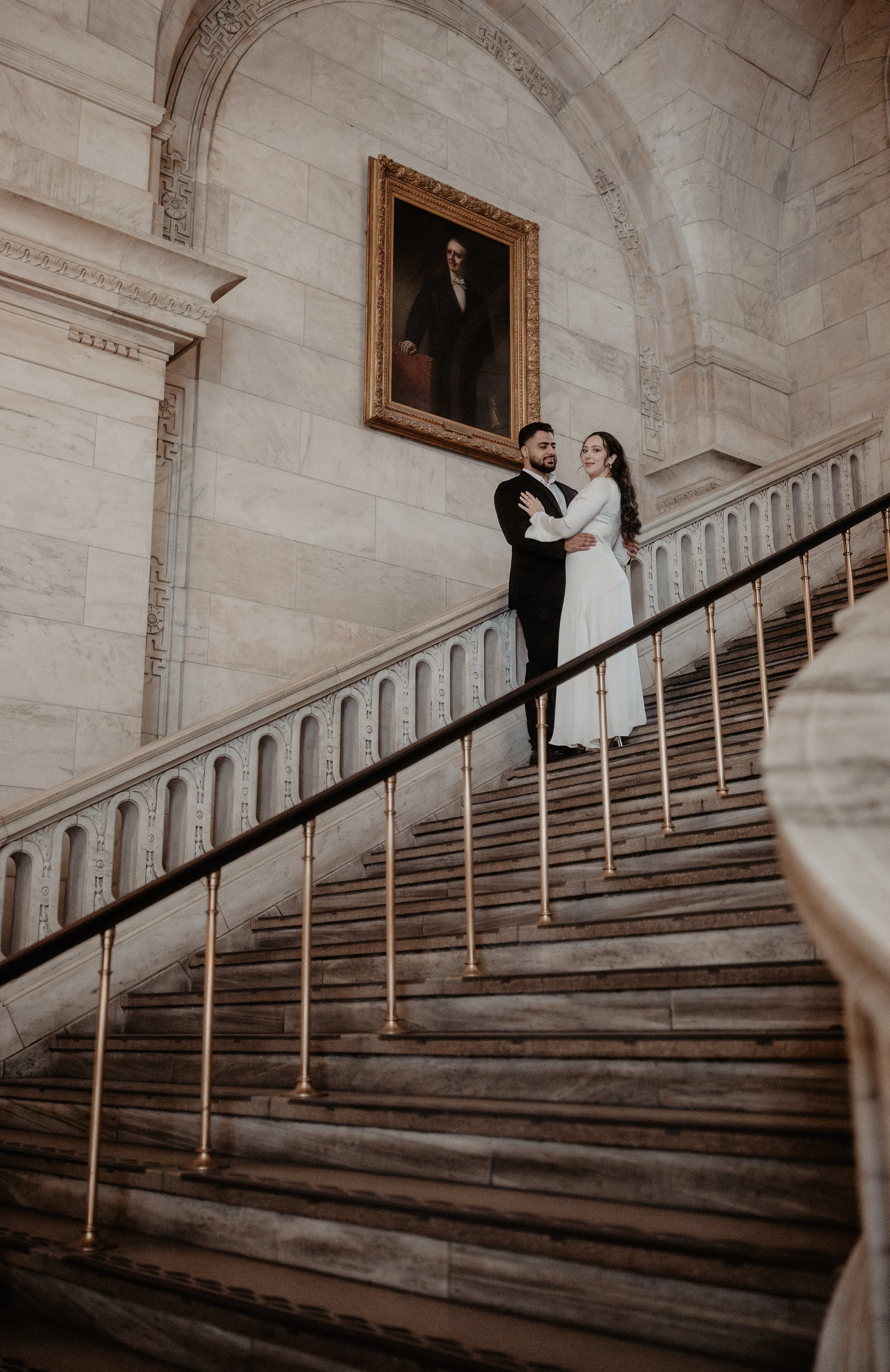 A couple standing on a grand staircase inside a historic building with marble walls, holding each other and looking at the camera. There is a large portrait of a man in a suit hanging on the wall behind them.