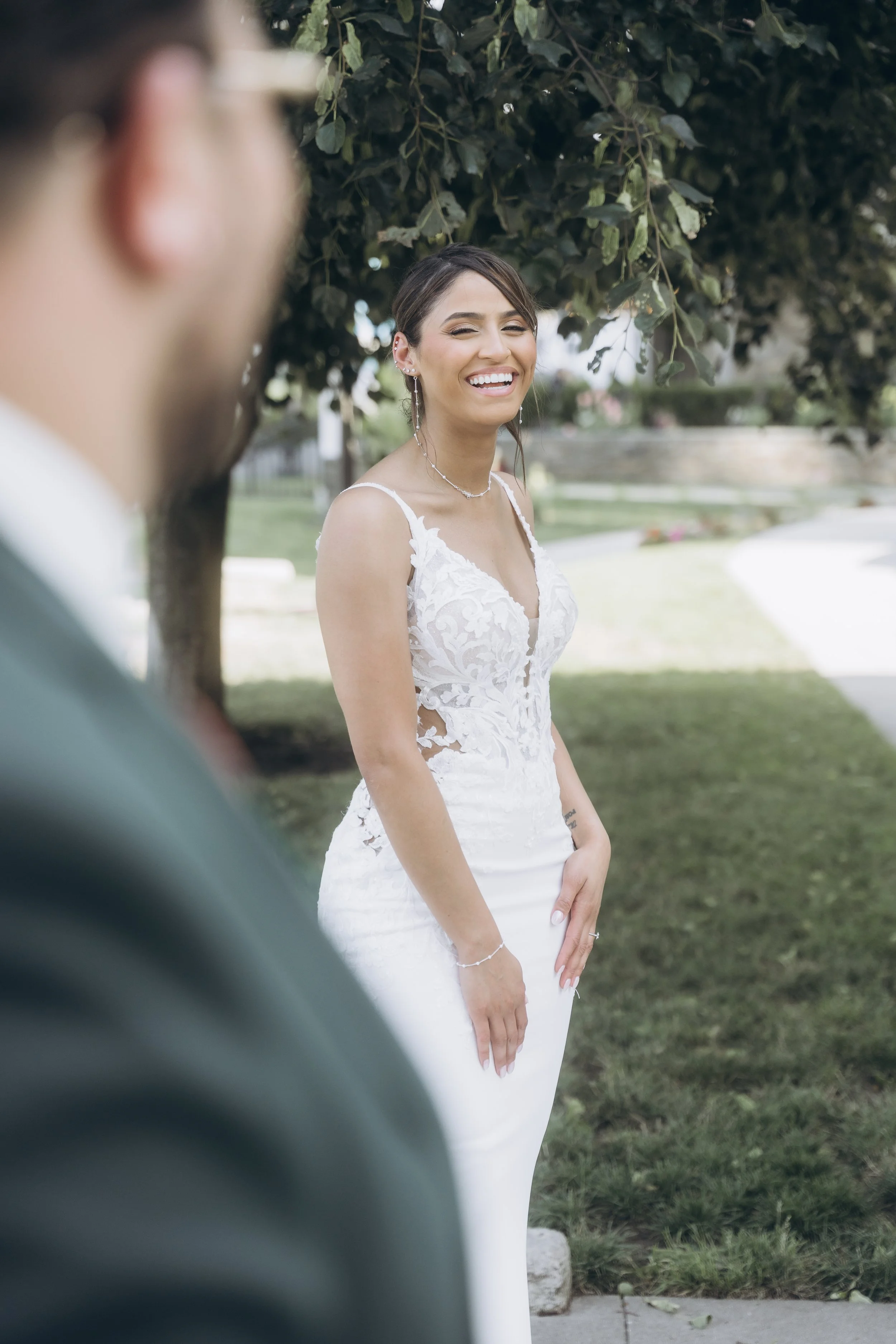 A bride in a white lace wedding dress smiling and looking at her groom, who is partially visible in the foreground wearing glasses and a green suit, outdoors under a tree.