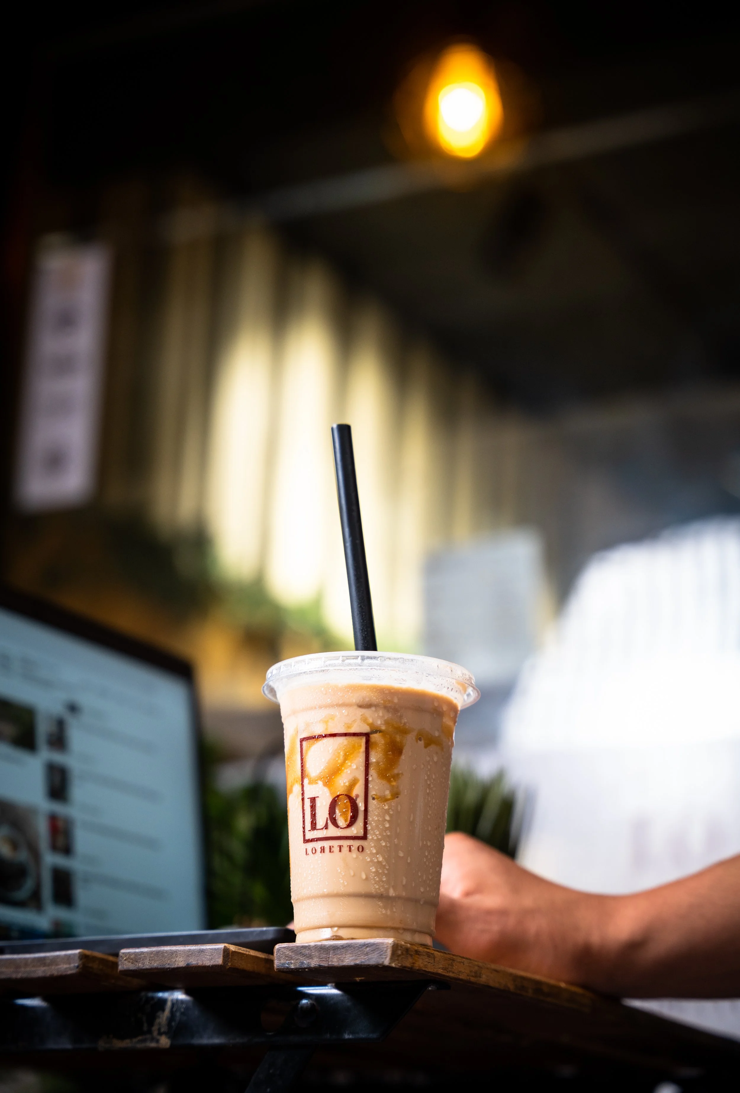 A cold iced coffee in a plastic cup with a black straw, placed on a wooden table with a person holding it. Background includes a blurred monitor, a yellowish light, and indoor decor.