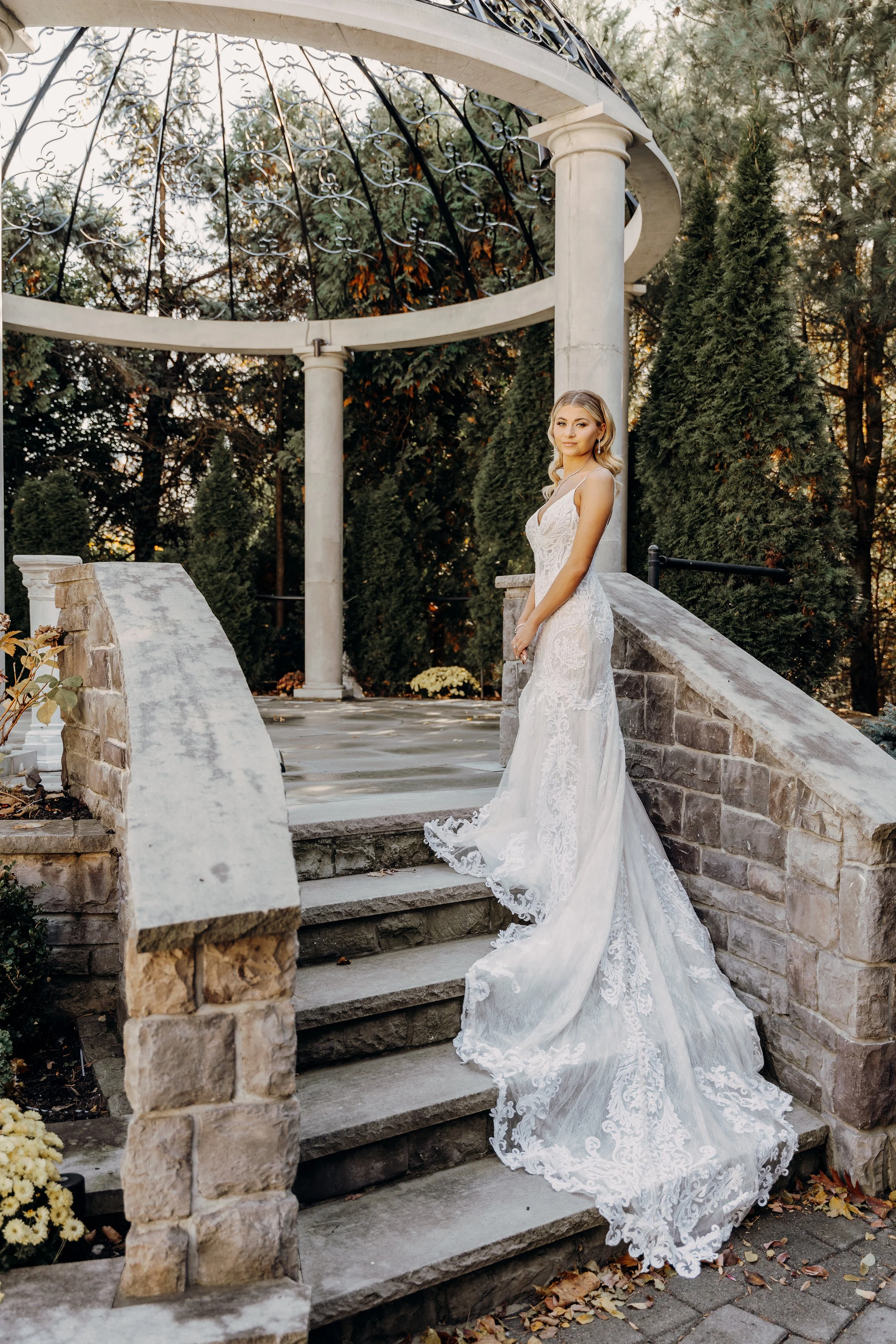 A woman in a white lace wedding gown stands on stone steps outdoors, with greenery and trees in the background.