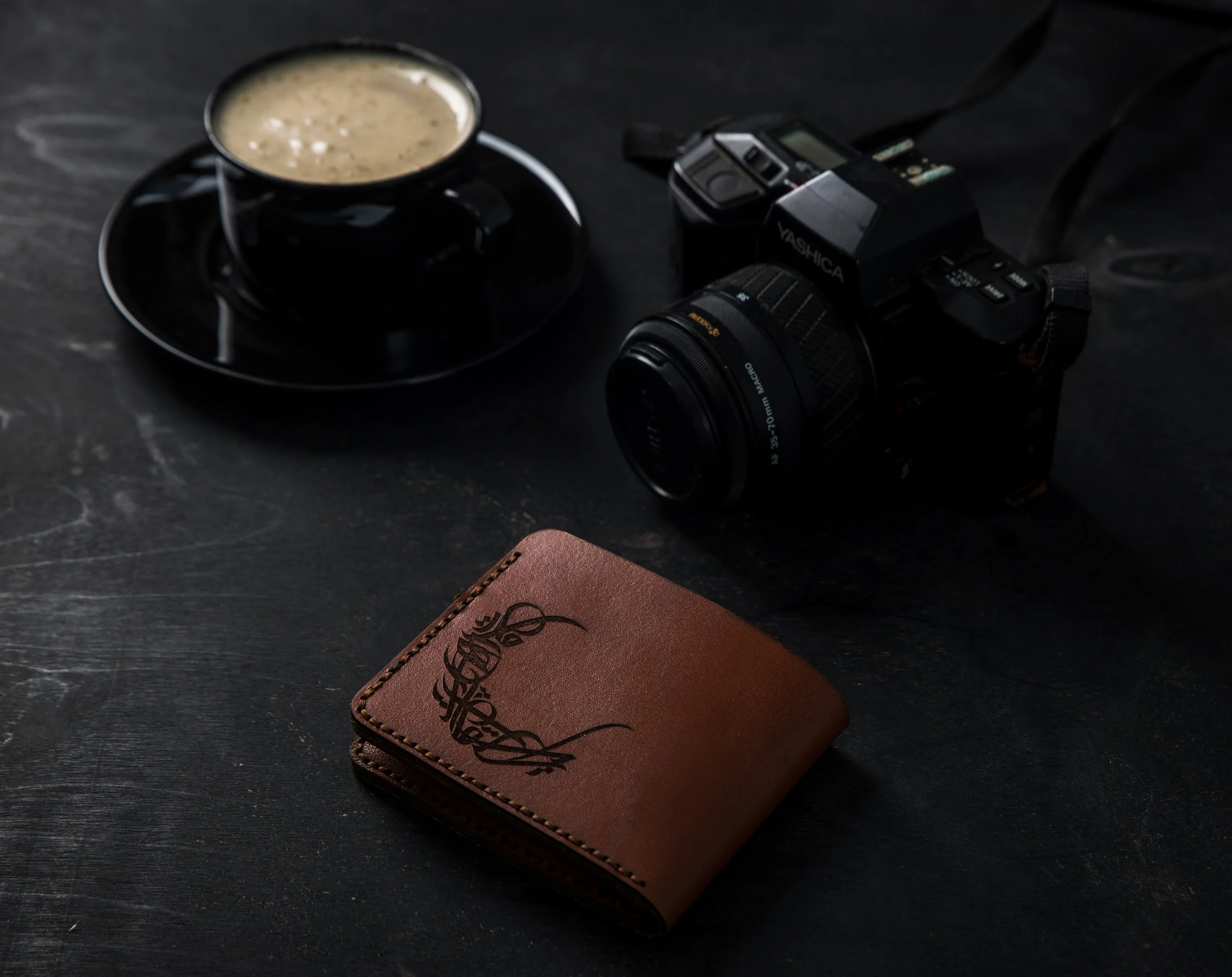 A black cup of coffee on a saucer, a camera, and a brown leather wallet on a dark surface.