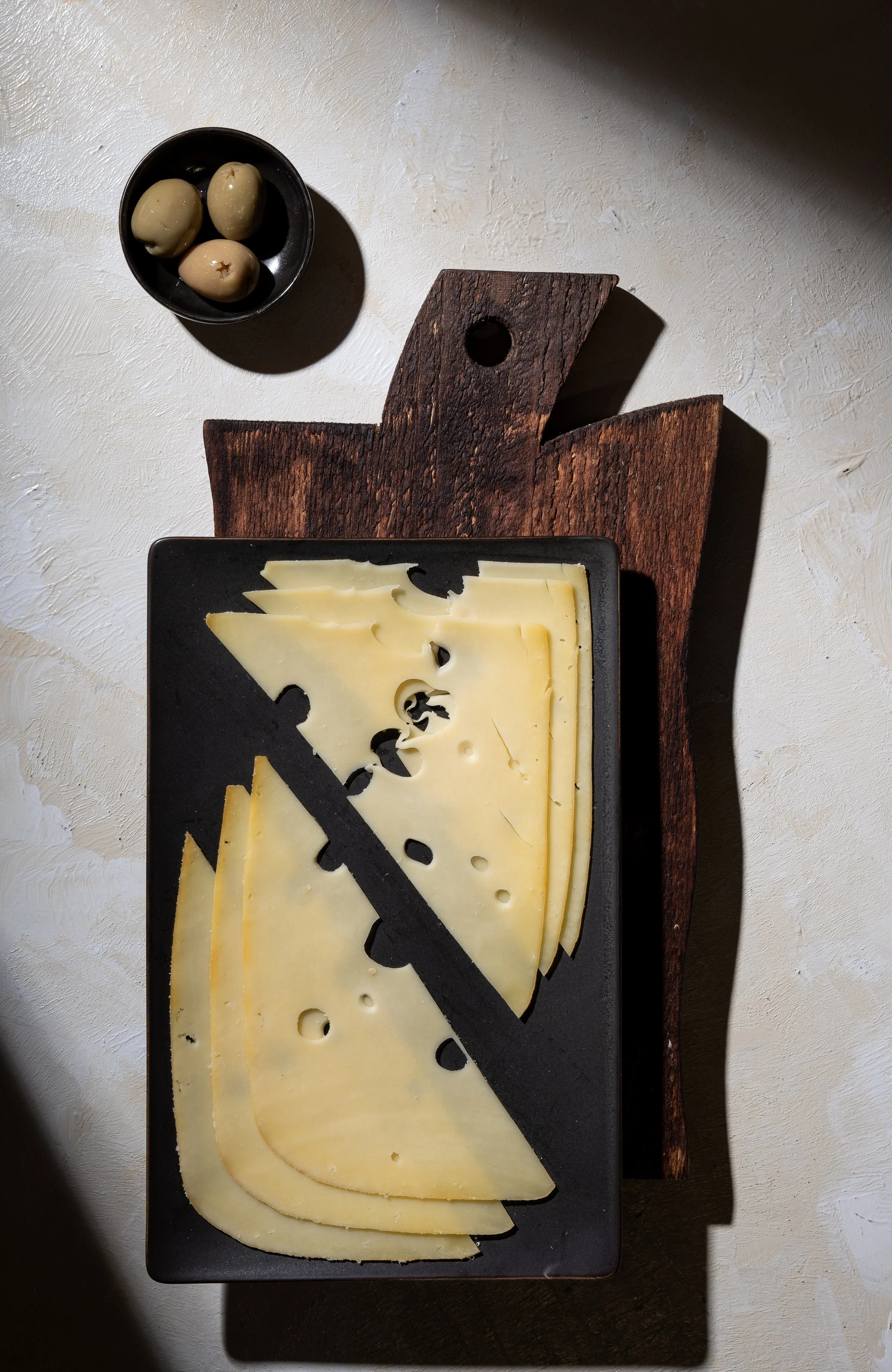 Sliced Swiss cheese on a black tray, placed on a rustic wooden cutting board, with a small black bowl of green olives nearby.