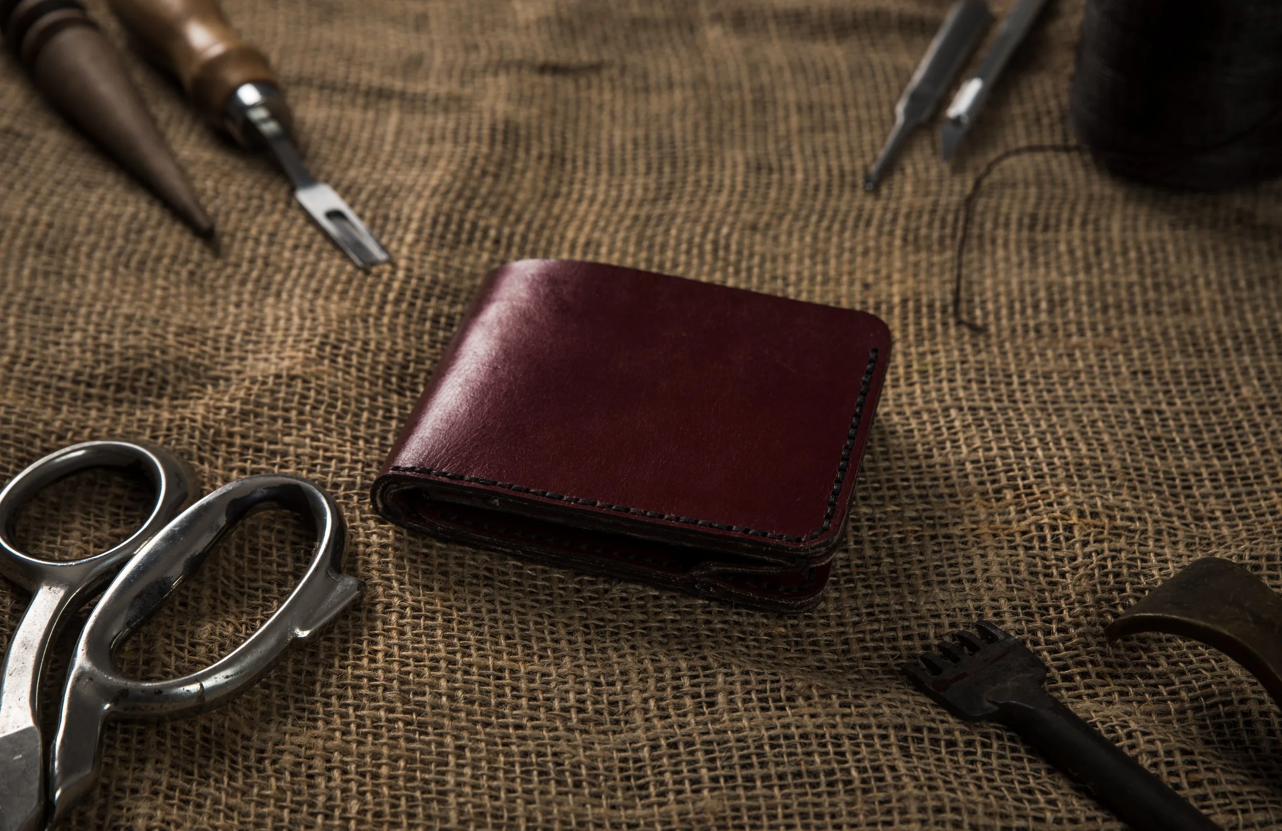 A folded red leather wallet on burlap fabric surrounded by leatherworking tools including scissors, a stamp, a carving tool, and a small fork.