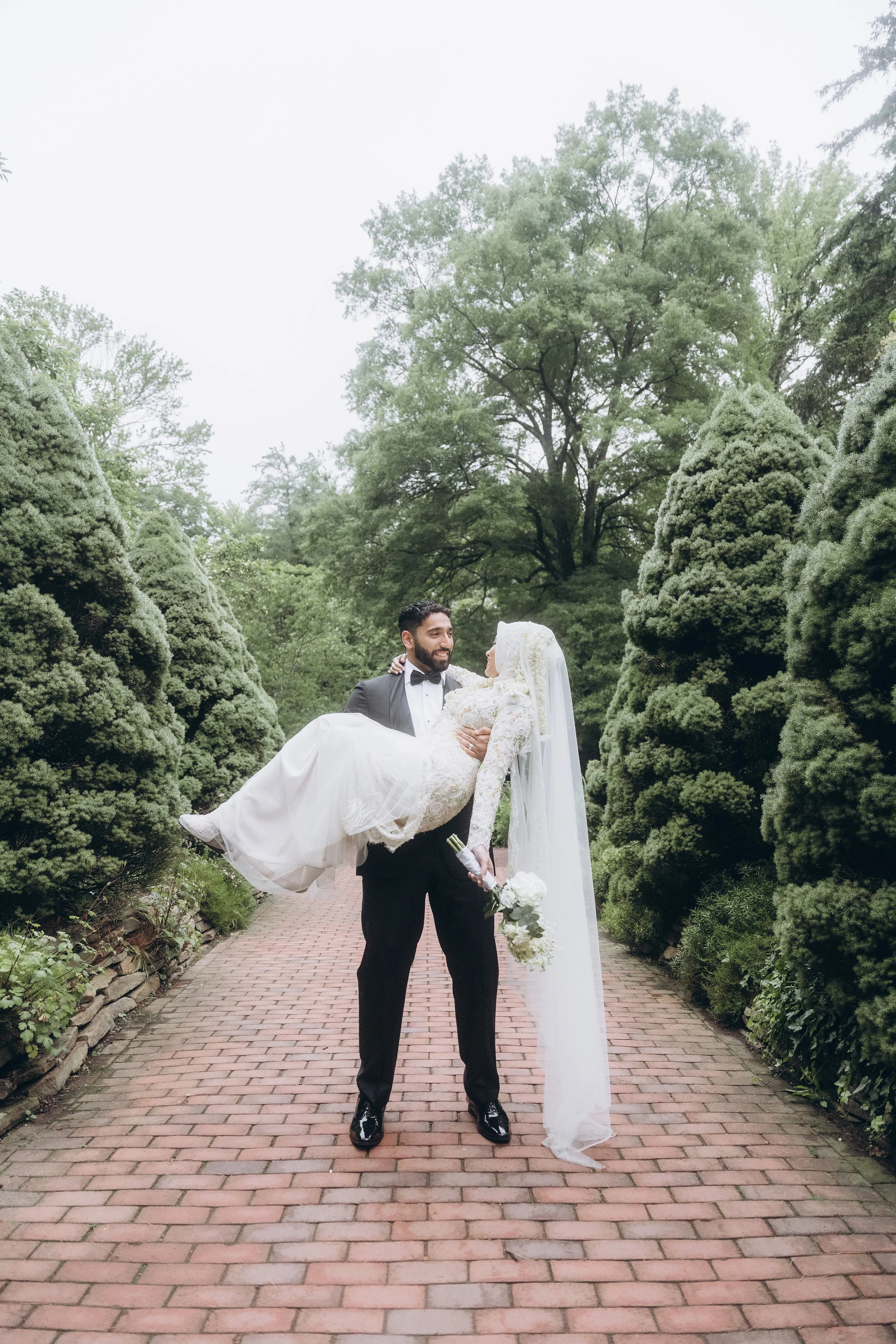 A groom in a tuxedo lifting a bride in a wedding dress holding a bouquet of flowers in a garden with greenery and trees.