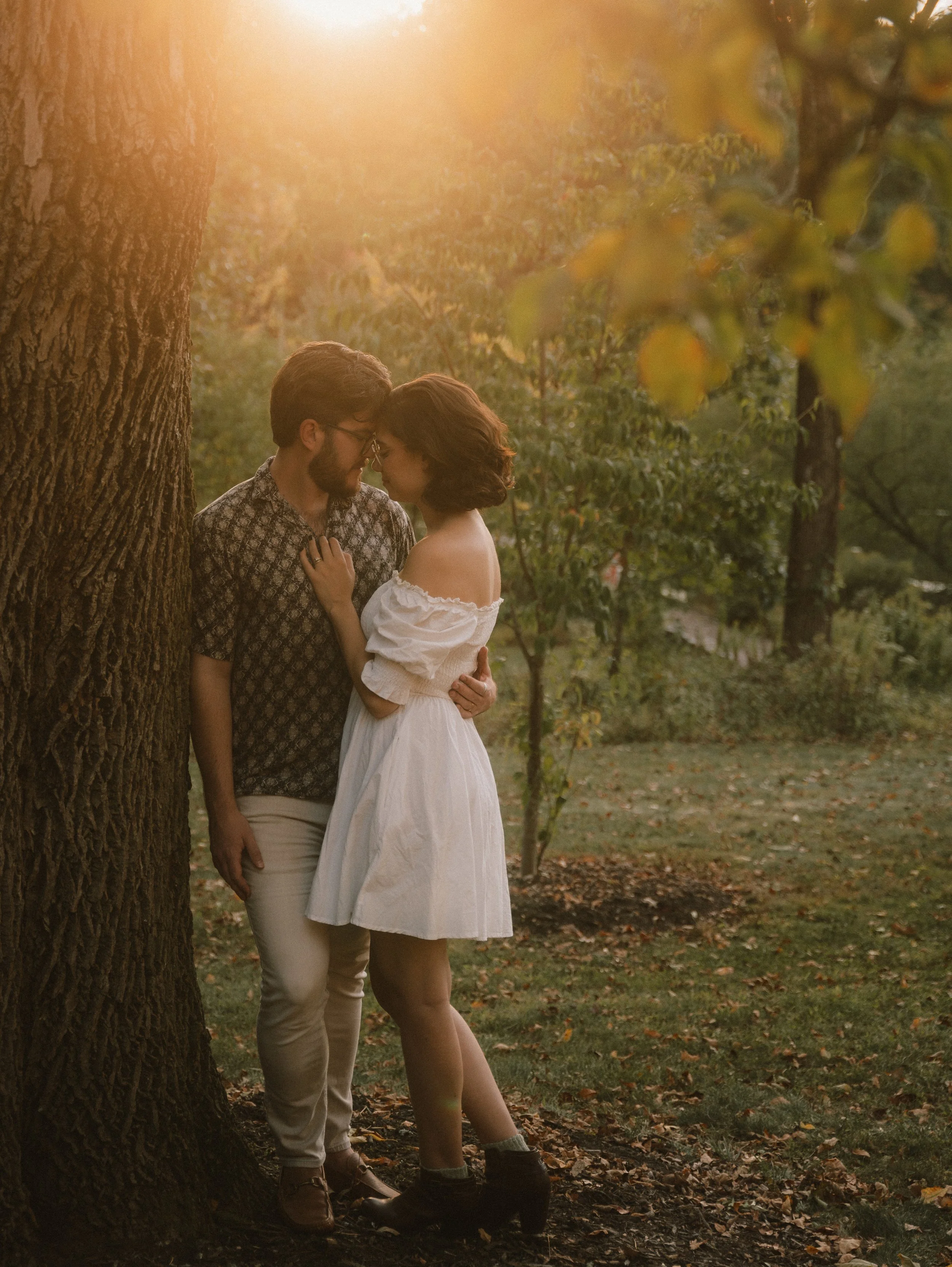 A couple stands close to each other under a large tree during sunset in a park, with their foreheads touching and eyes closed.