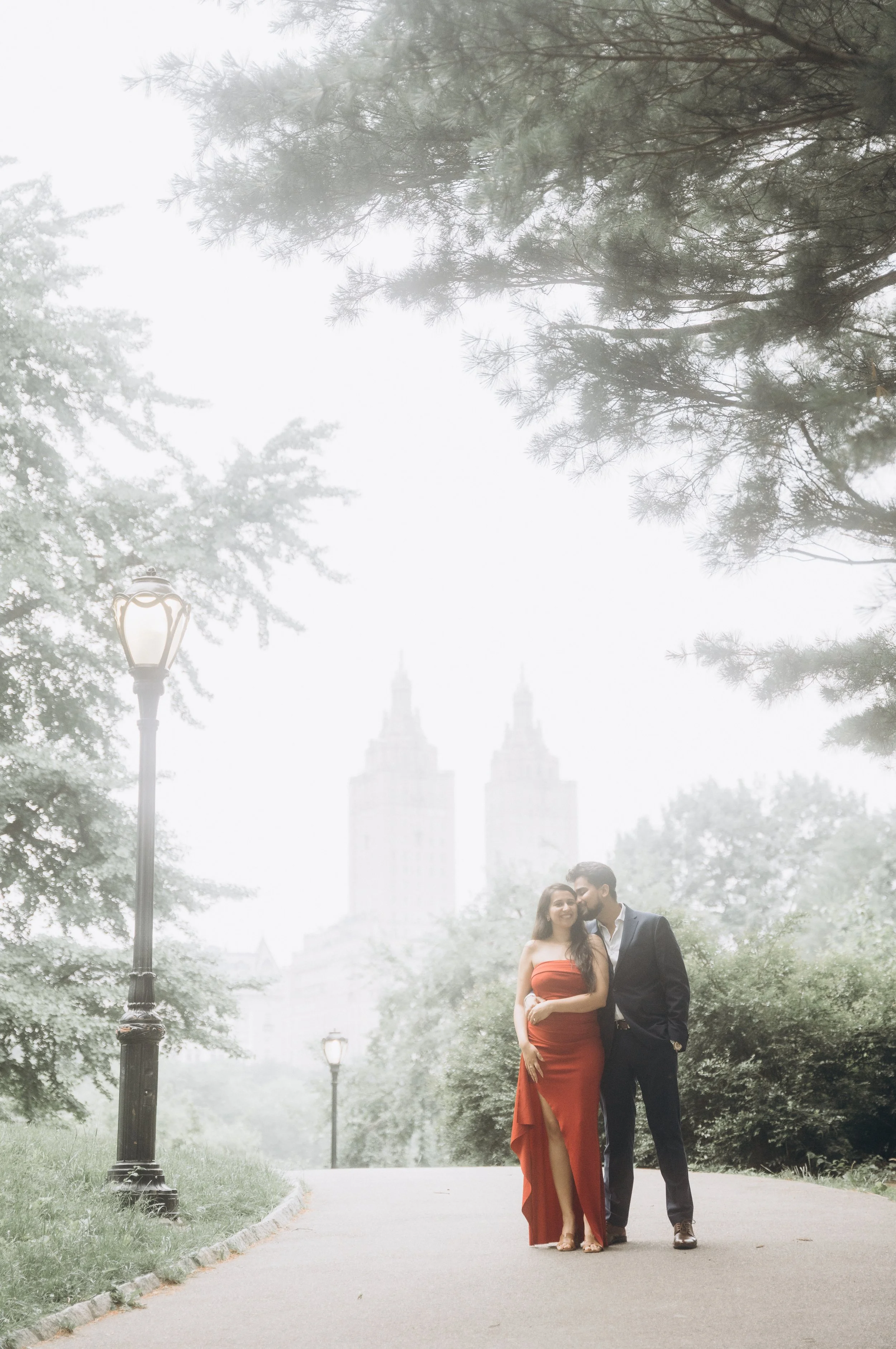 A couple walking on a park pathway with trees and street lamps, with tall buildings in the background, under foggy weather.