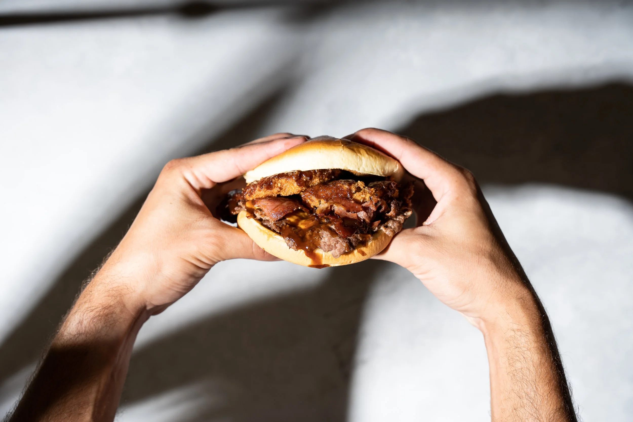 Person holding a sandwich with bacon, fried chicken, and barbecue sauce in a bun.